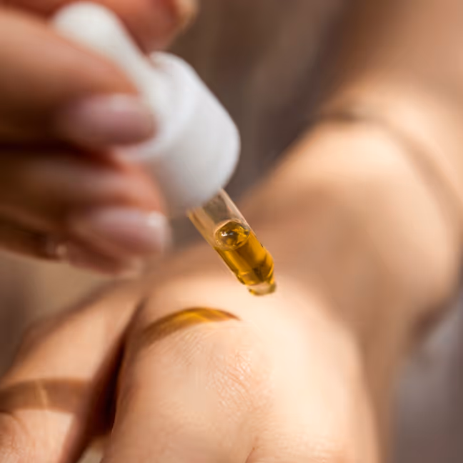 Close-up of a hand holding a dropper applying golden serum onto the back of another hand.