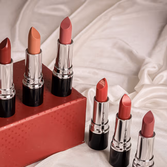 Six tubes of lipstick in various shades of red and pink arranged with three on a red box and three on white fabric.