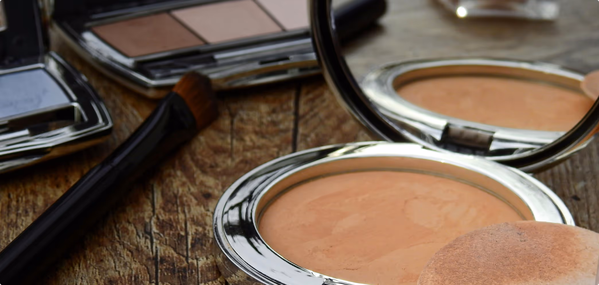 Compact powder with mirror and makeup brush on a wooden surface with an eyeshadow palette in the background.