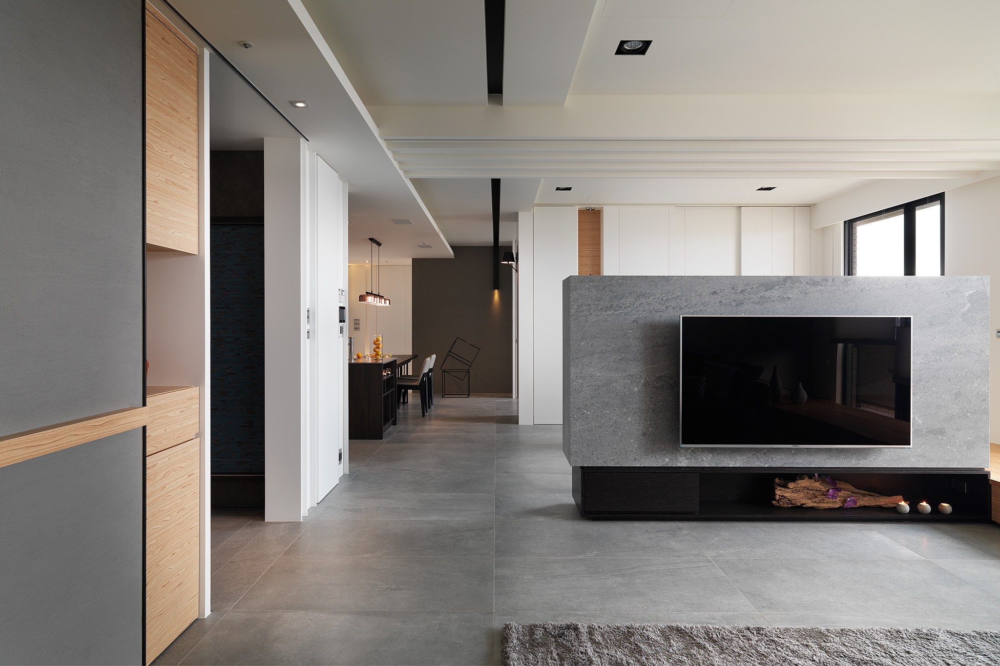 Modern open living space with a mounted flat-screen TV on a gray stone partition and a dining area with pendant lights in the background.