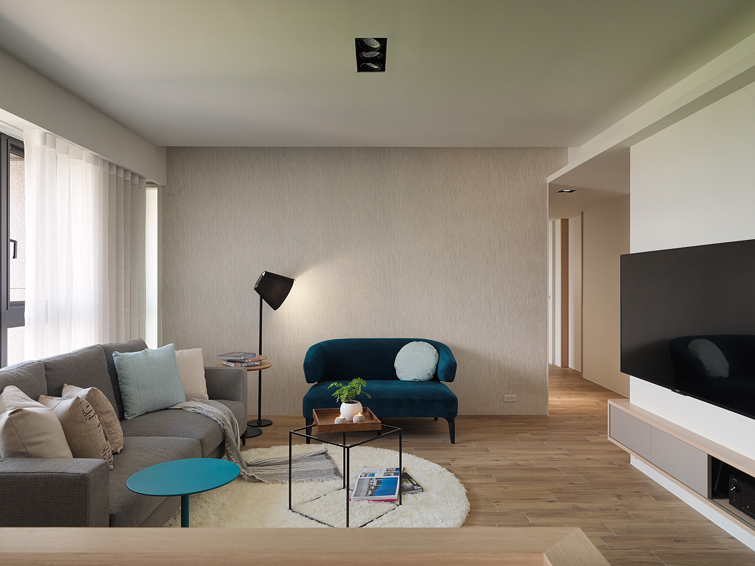 Minimalist living room with L-shaped gray sofa, glass coffee table, large windows with blinds, and modern art pieces.