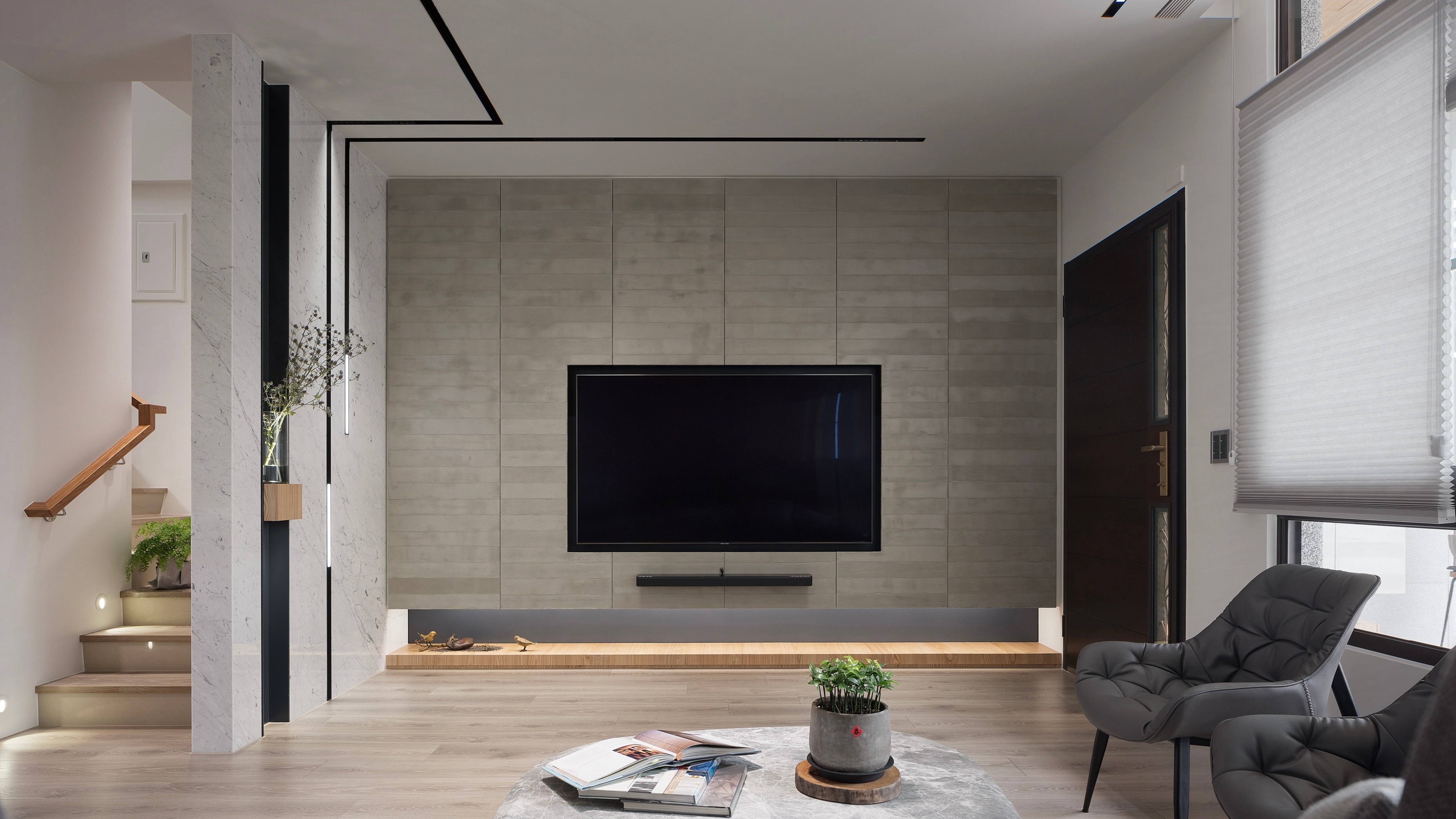 Modern living room with a wall-mounted flat-screen TV, two gray chairs, a round marble coffee table with a plant and books, and a staircase with wooden handrail.