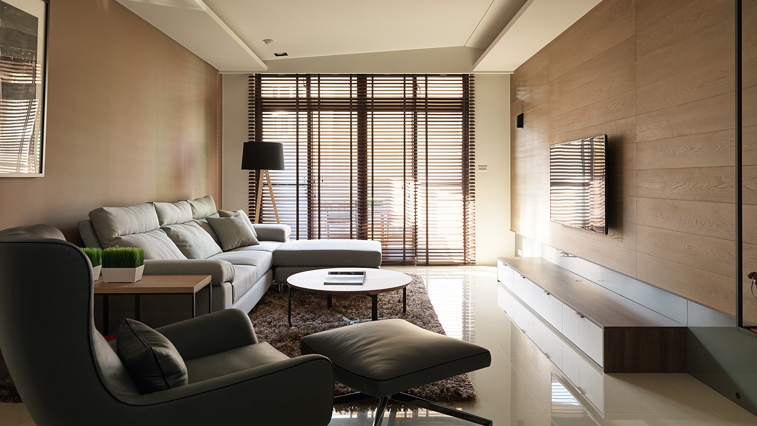 Modern living room with a gray sectional sofa, black armchair with ottoman, round coffee table, floor lamp, and a wall-mounted TV on a wood-paneled wall with large window blinds.