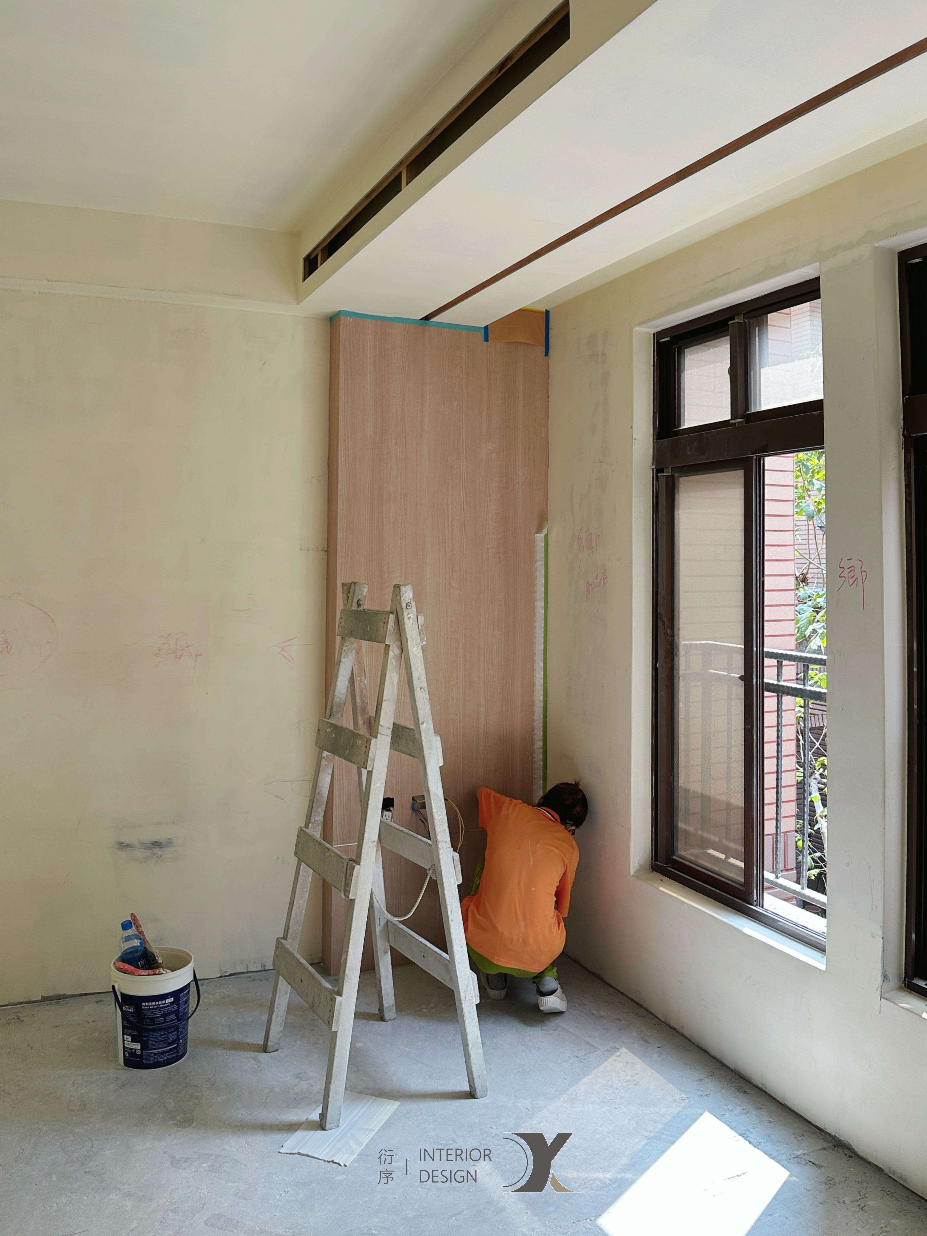 Two construction workers in hard hats standing on ladders working near a large window in a room under renovation with sunlight streaming in.