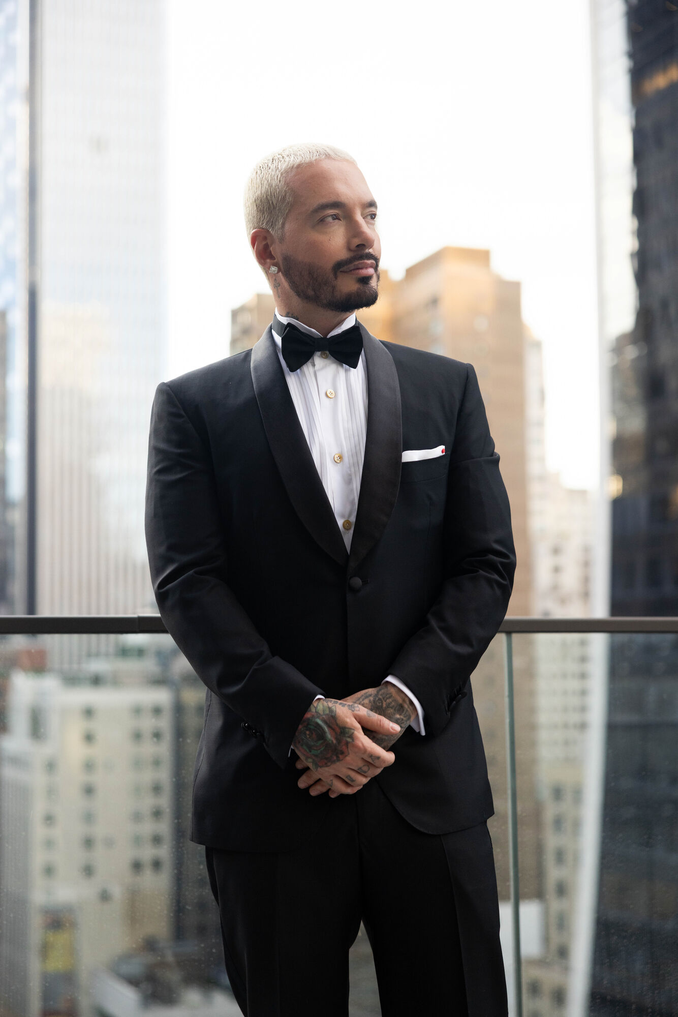 J Balvin in a black tuxedo and bow tie against floor-to-ceiling windows over the city.