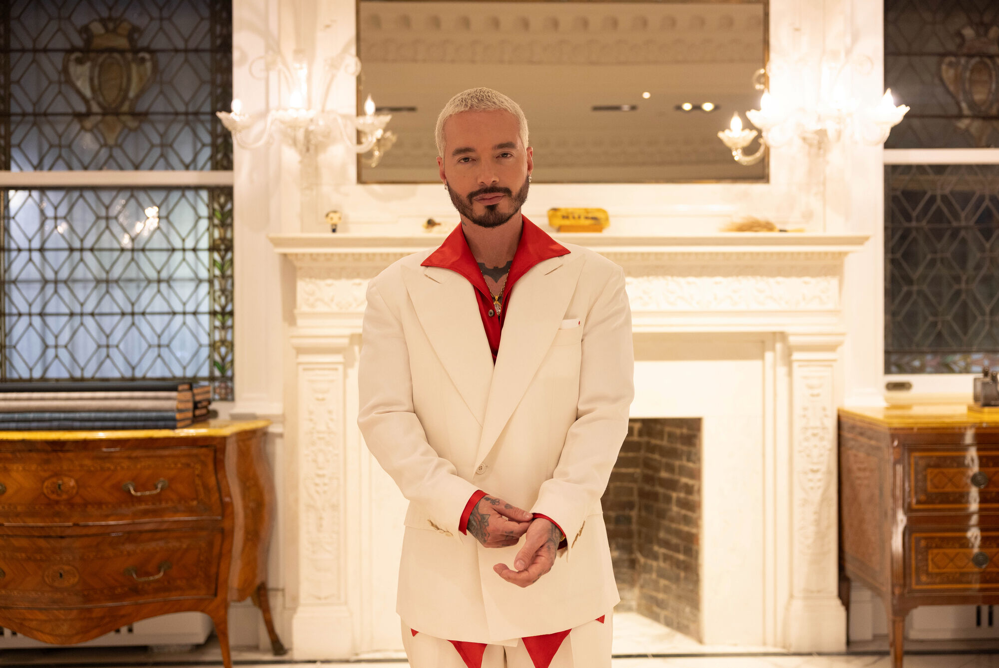 J Balvin in a white Kiton suit with a red shirt in front of a marble fireplace.