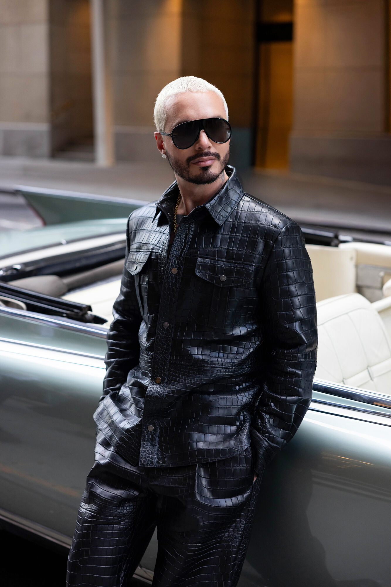 J Balvin in a black crocodile-texture jacket and sunglasses beside a vintage convertible.