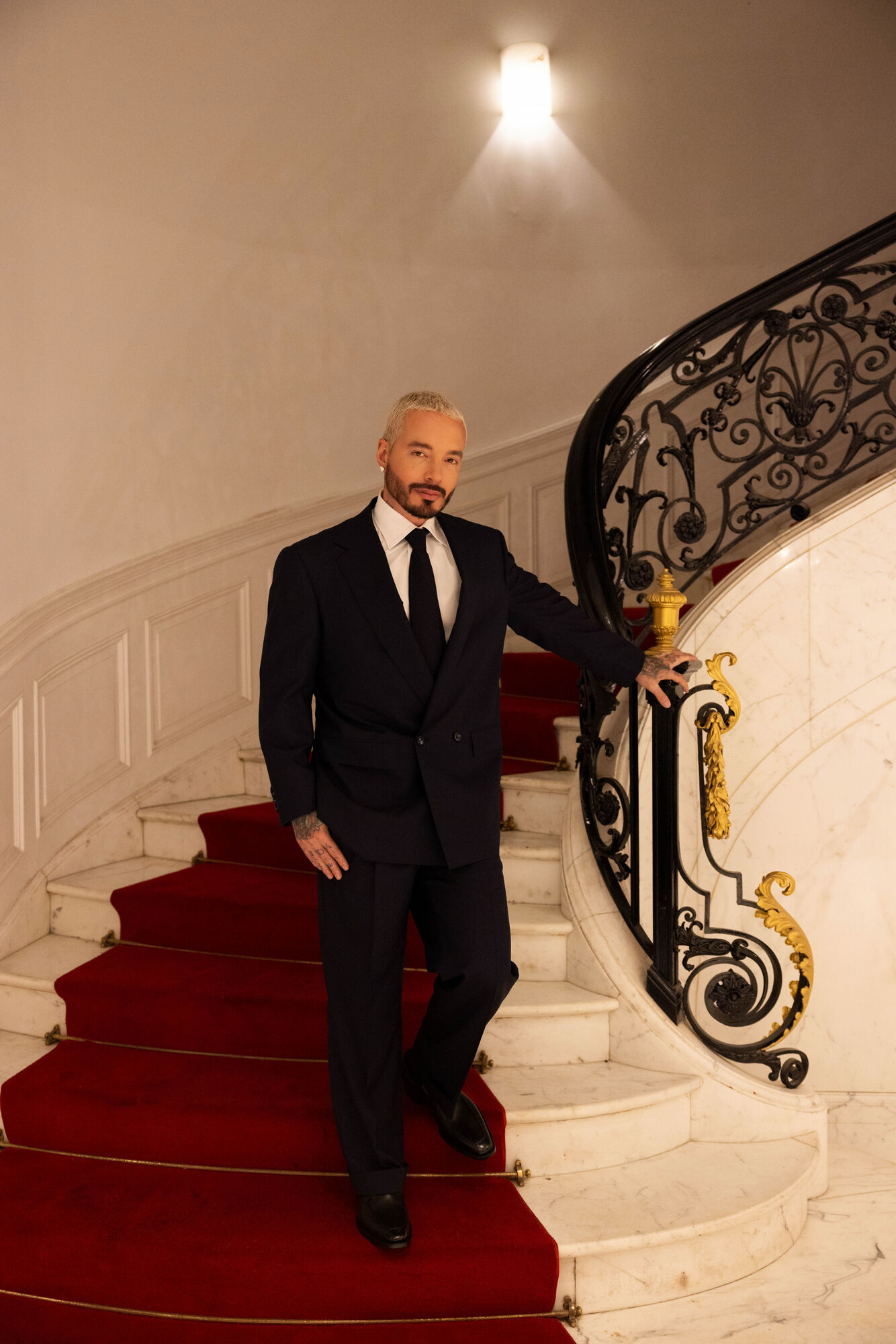 J Balvin in a dark double-breasted suit on a curved marble staircase with a red runner.