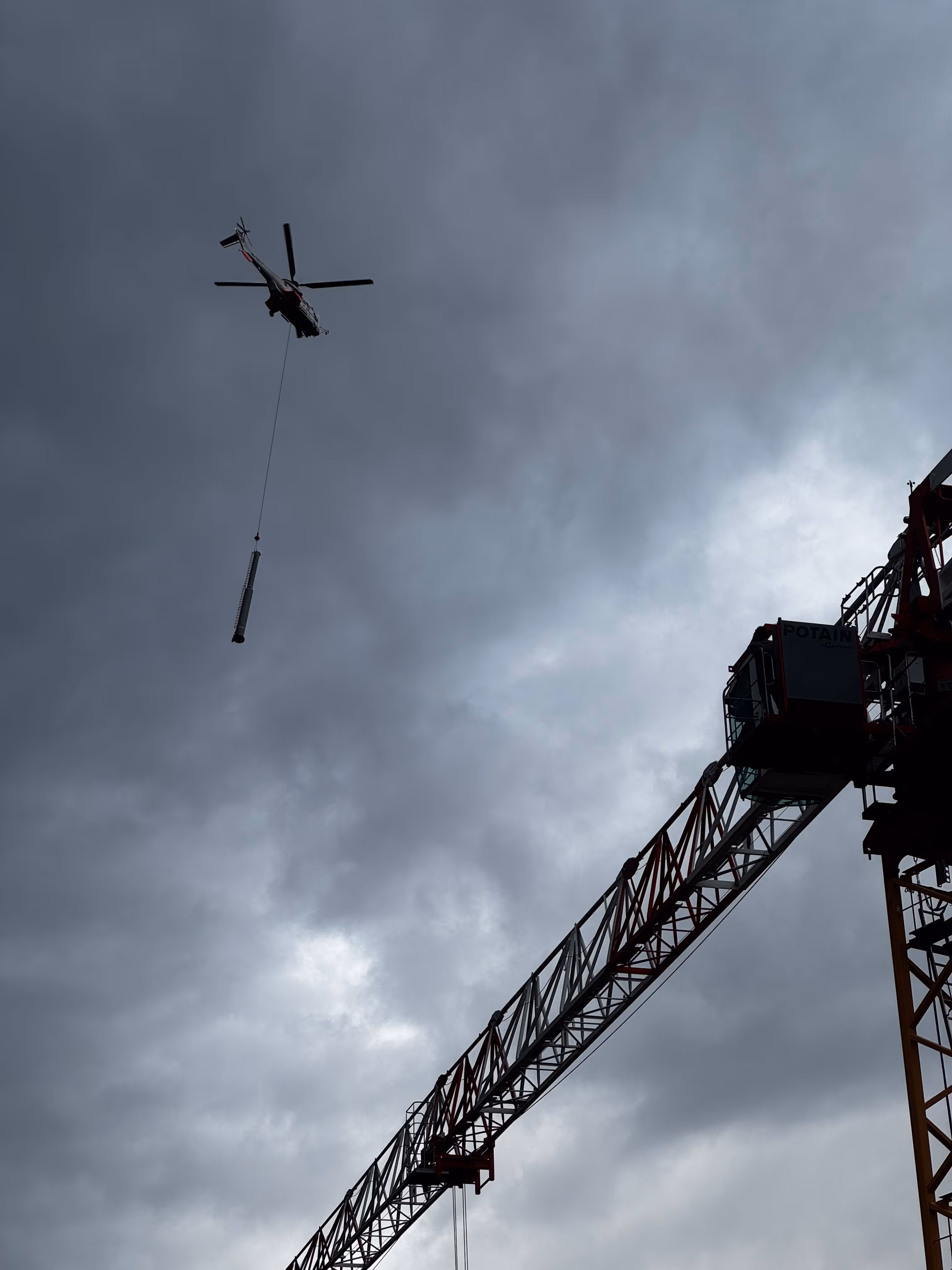 Hélicoptère transportant une charge suspendue au-dessus d'une grue contre un ciel nuageux.