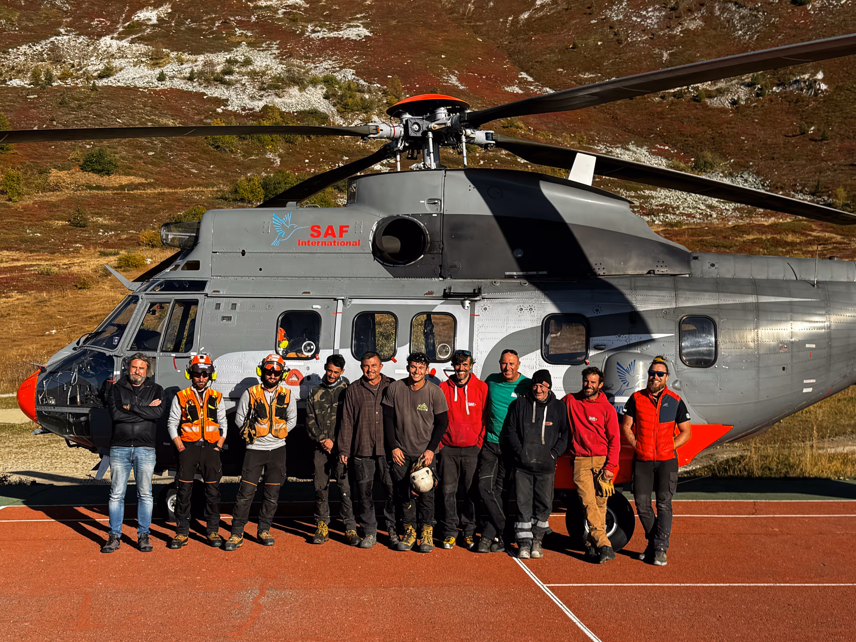 Une équipe de onze hommes debout devant un hélicoptère gris et rouge marqué SAF International, posant sur une surface rouge avec un paysage montagneux en arrière-plan.