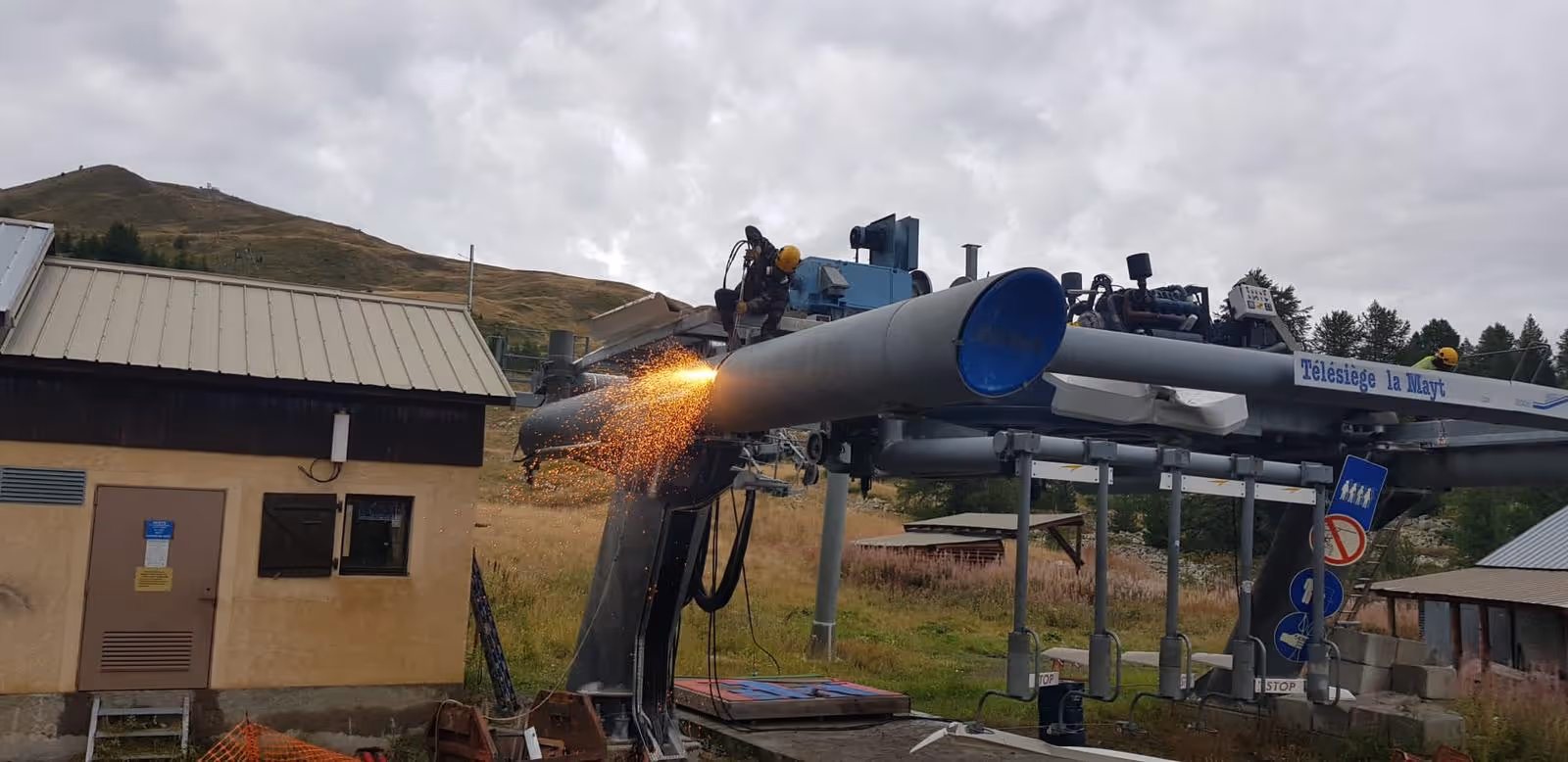 Un ouvrier avec un casque jaune effectuant une soudure sur une structure métallique d'un télésiège en montagne par temps nuageux.