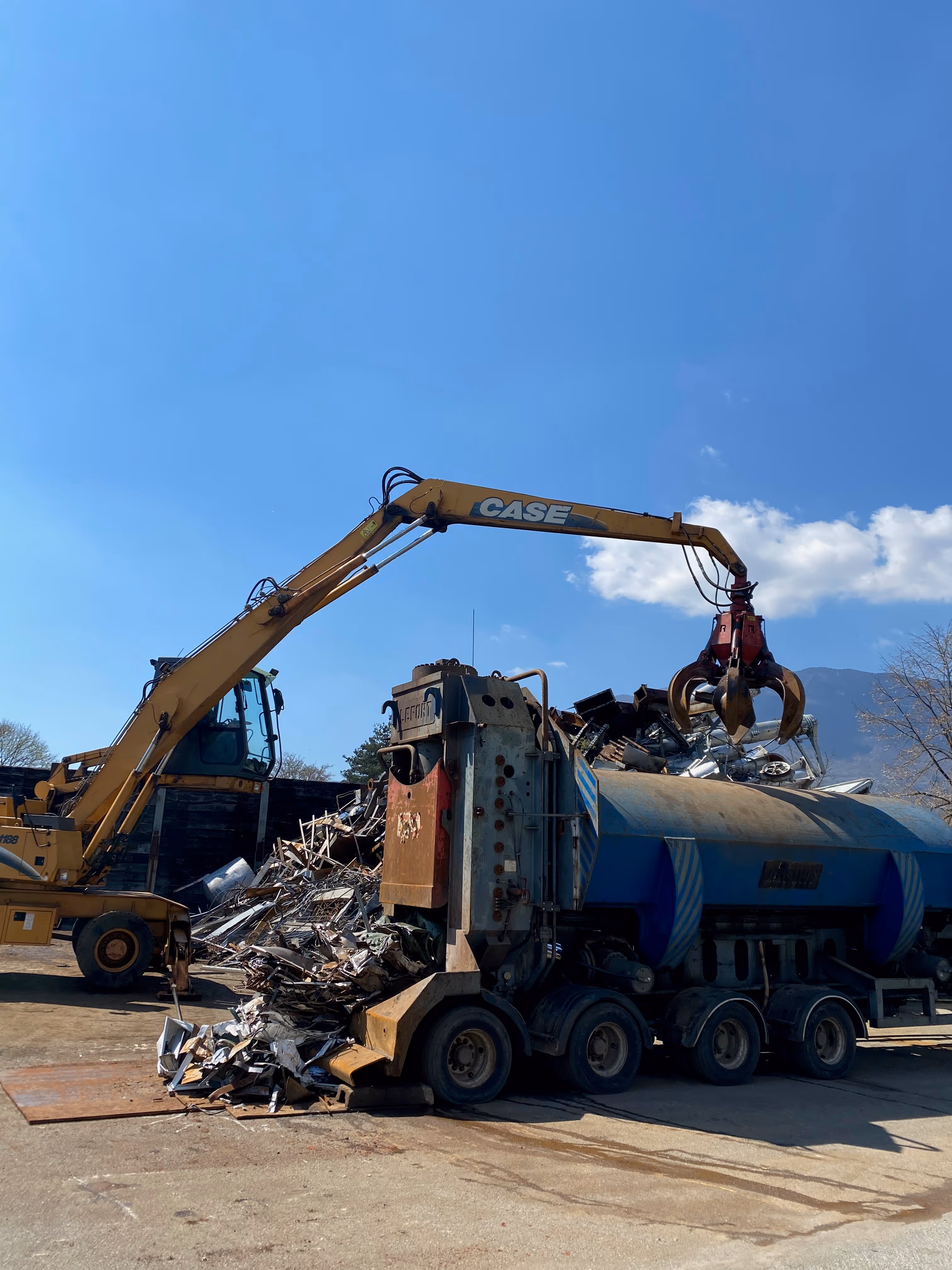 Une pelleteuse jaune CASE équipée d'une pince saisit des déchets métalliques pour les charger dans un camion bleu volumineux.