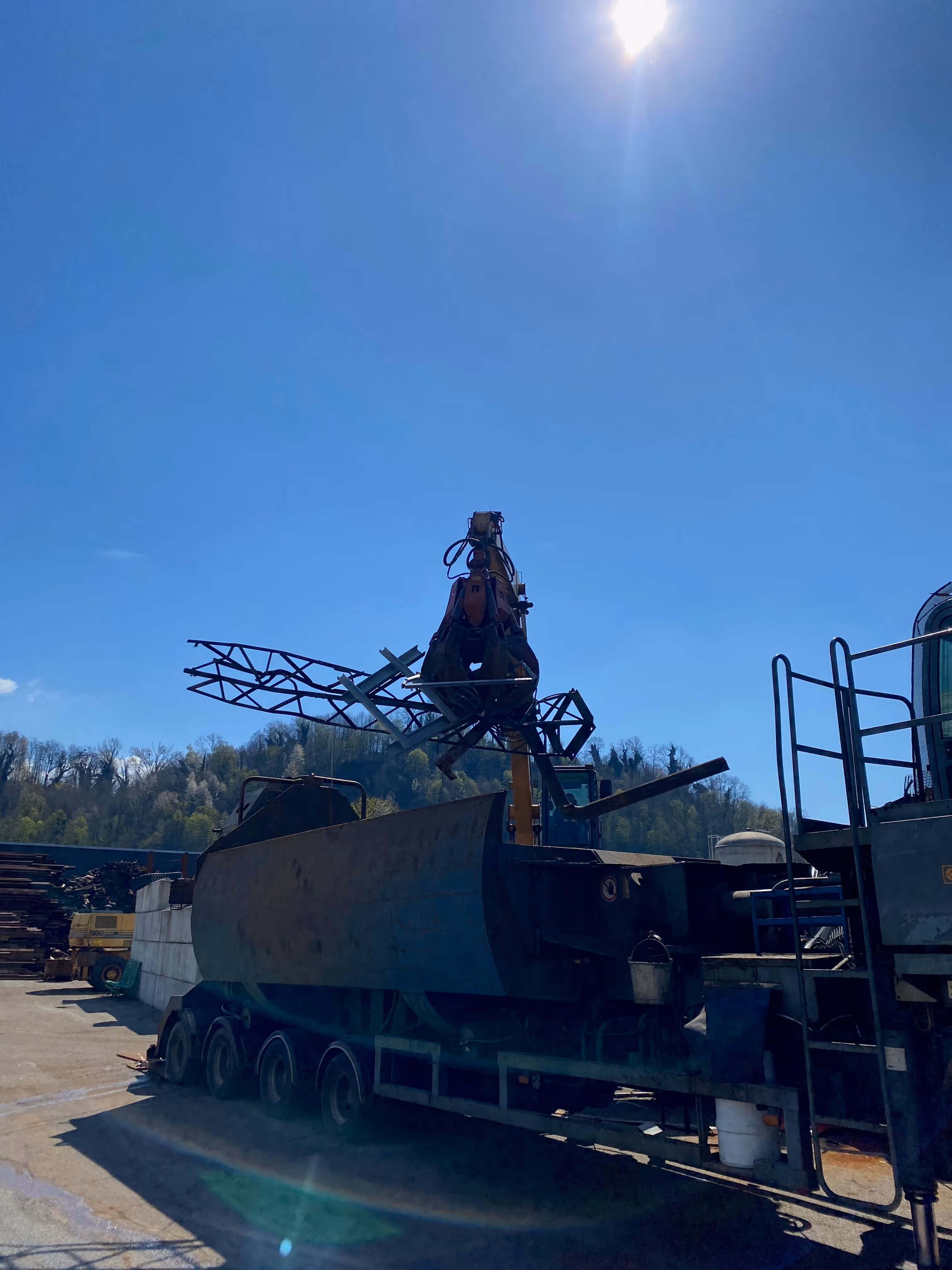 Camion lourd équipé d'une grue mécanique soulevant une structure métallique triangulée sur fond de ciel bleu clair et soleil brillant.