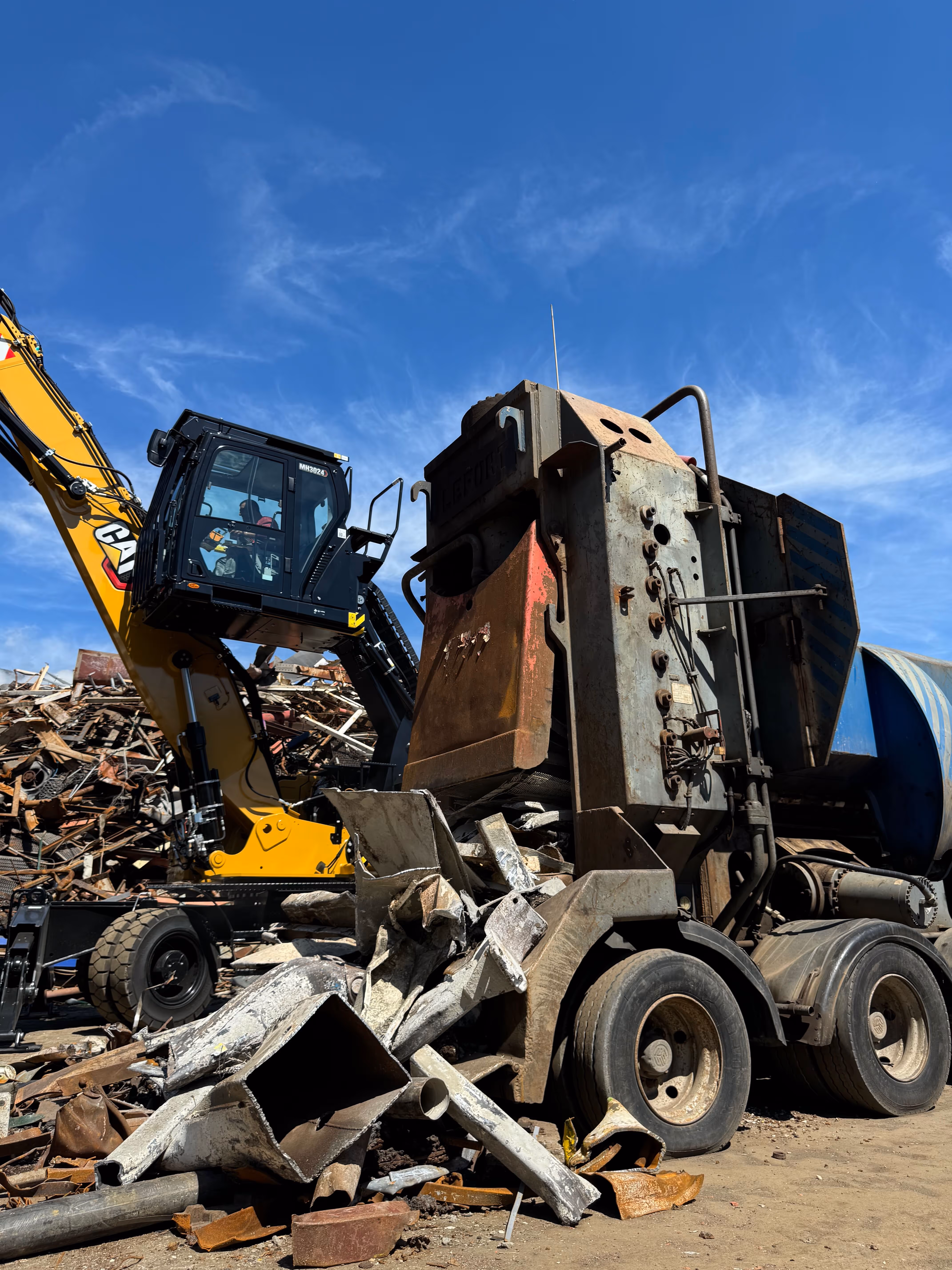 Vieux camion malaxeur à béton rouillé parmi des débris métalliques avec une pelle mécanique jaune en arrière-plan sous un ciel bleu.