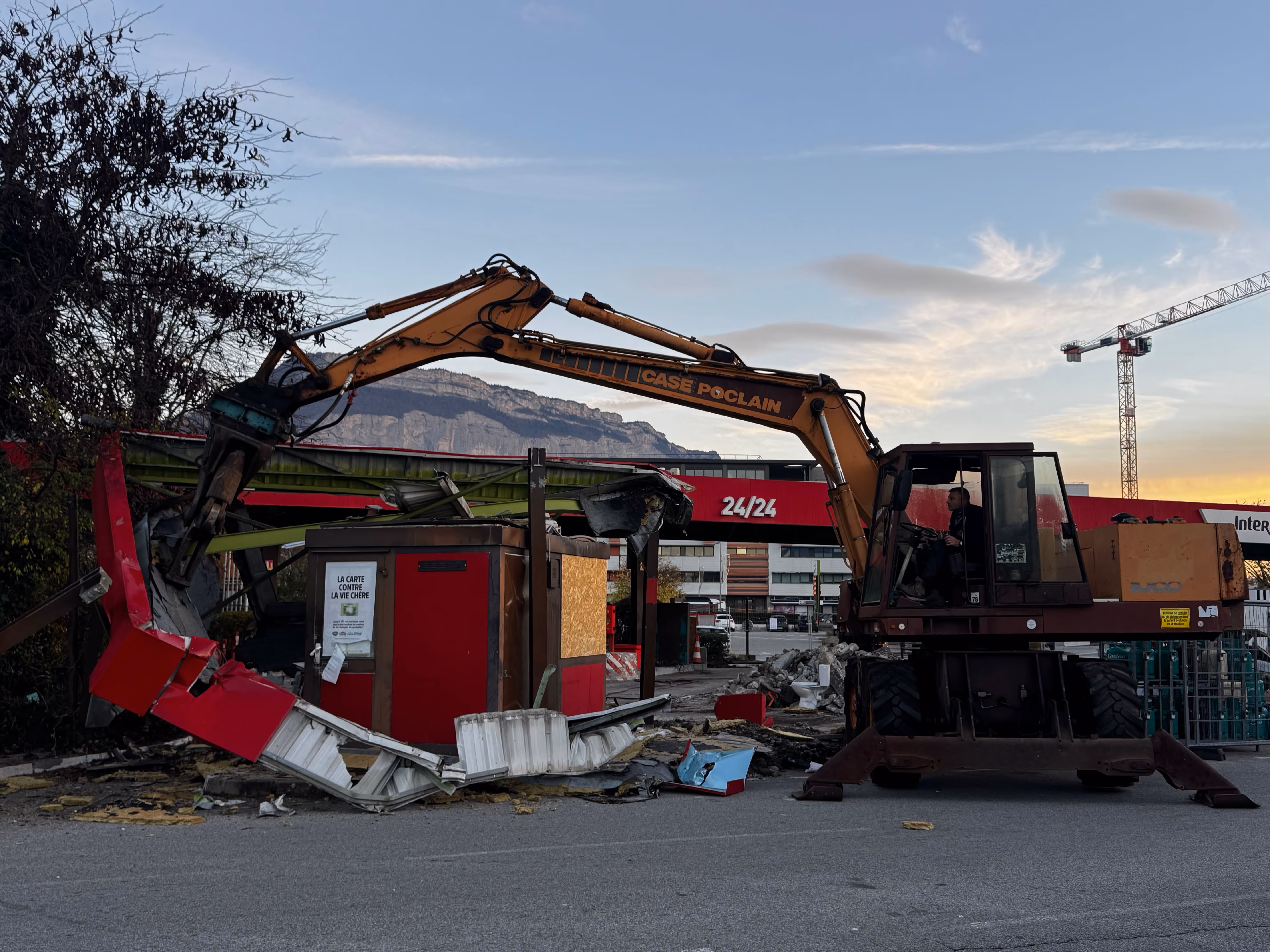 Pelle de démolition Case Poclain orange détruisant une structure métallique rouge sur un chantier, avec une station-service Intermarché 24/24 et une montagne en arrière-plan au crépuscule.