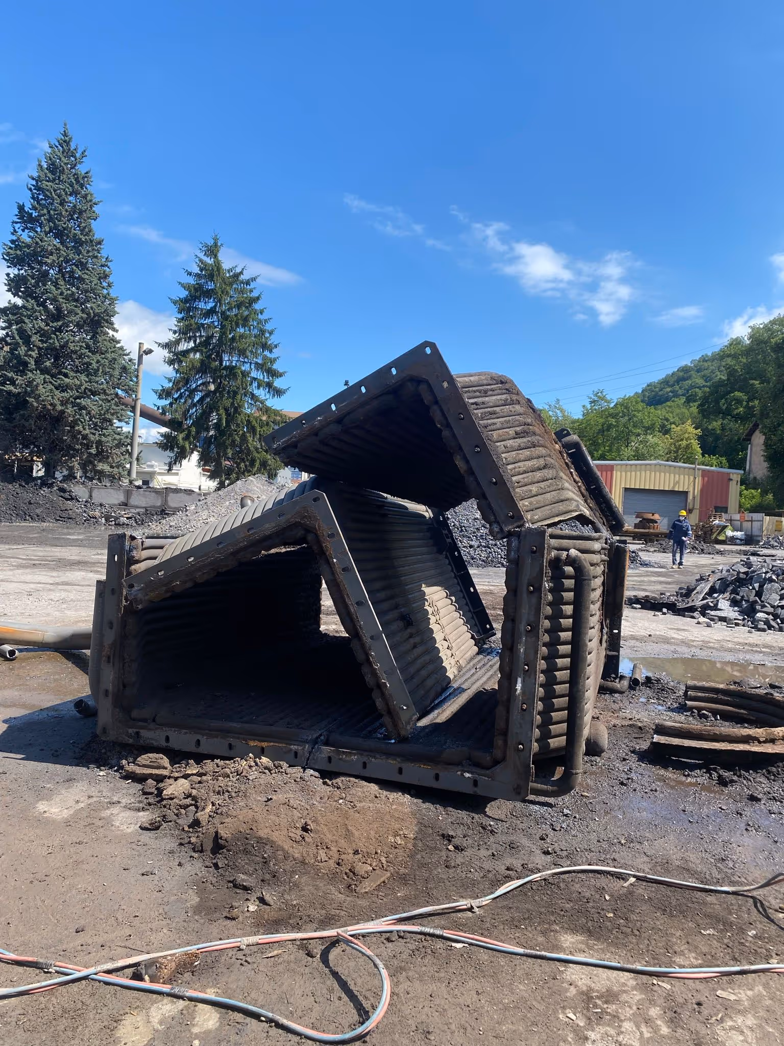 Grande structure métallique industrielle en tubes d'acier, partiellement découpée et empilée sur un terrain de chantier poussiéreux sous un ciel bleu dégagé
