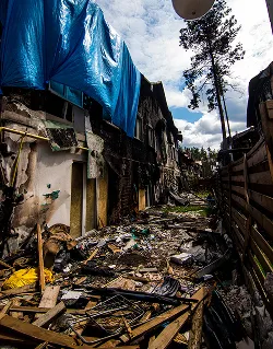 Verval en rommel in een nauwe steeg met oude gebouwen, blauw zeildoek en een hoge boom onder een bewolkte hemel