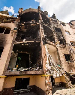 Beschadigd appartementengebouw met ingestorte balkons en verbrande gevel onder een blauwe lucht met wolken.