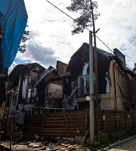 Brand beschadigde huizen met verbrande muren en puin rondom, onder een bewolkte hemel.