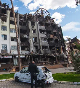 Man staat bij een zwaar beschadigde auto voor een verwoest appartementencomplex met een zwartgeblakerde gevel en instortingsgevaar.