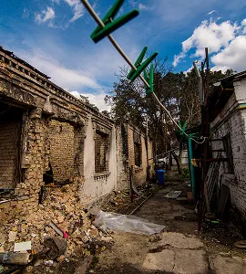 Verwoeste bakstenen muur van een gebouw met puin op de grond onder een blauwe lucht met wolken.