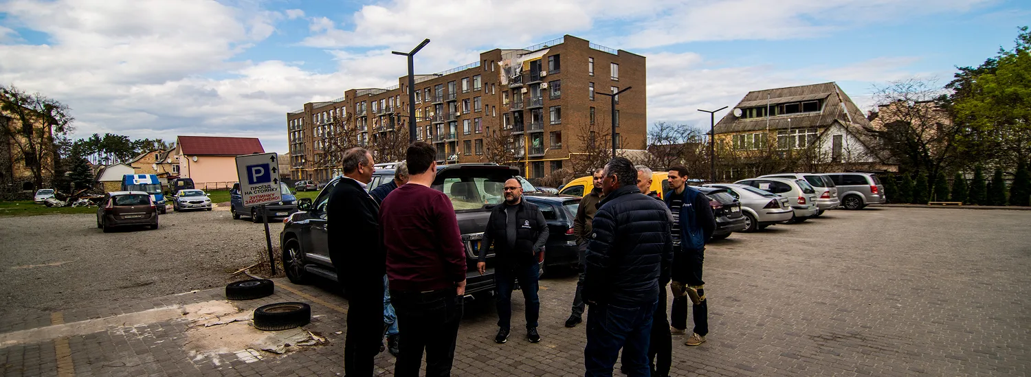 Een groep mannen die buiten staan bij een parkeerplaats met verschillende geparkeerde auto's en een achtergrond van appartement- en woonhuizen.