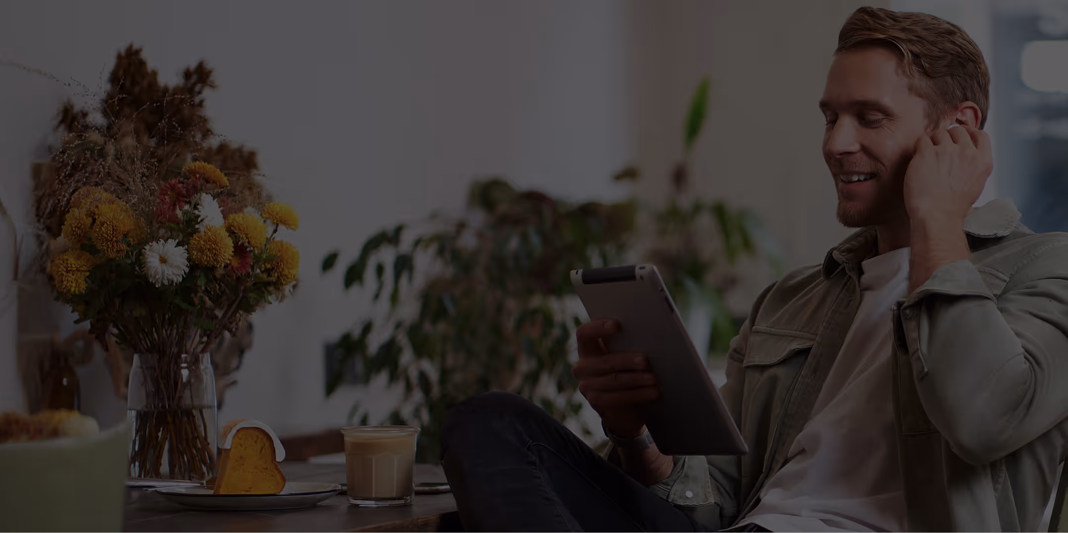 Smiling man wearing wireless earbuds and holding a tablet, sitting in a cozy room with flowers, coffee, and green plants.