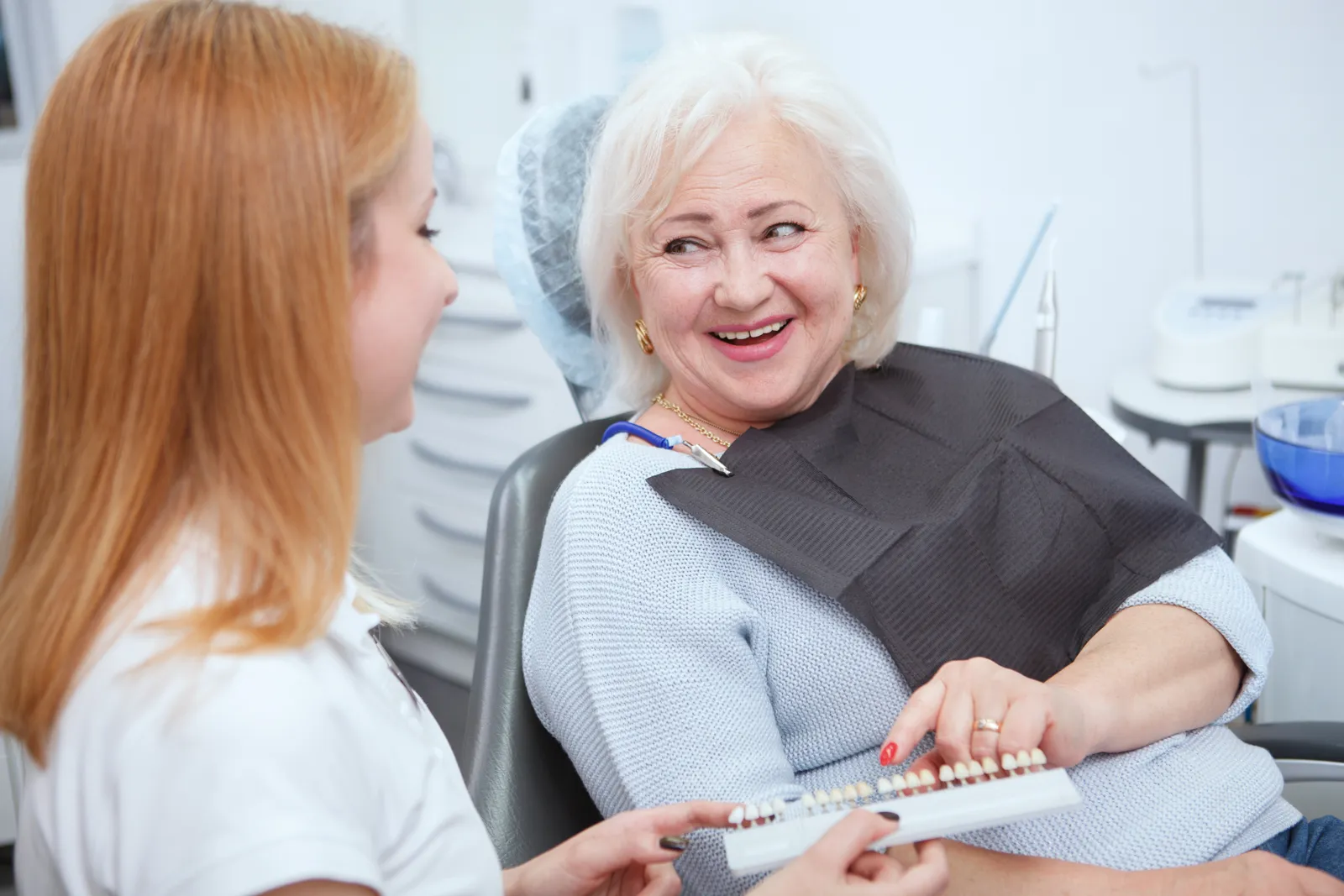 Older woman smiling in the dental chair while being shown a tooth shade guide