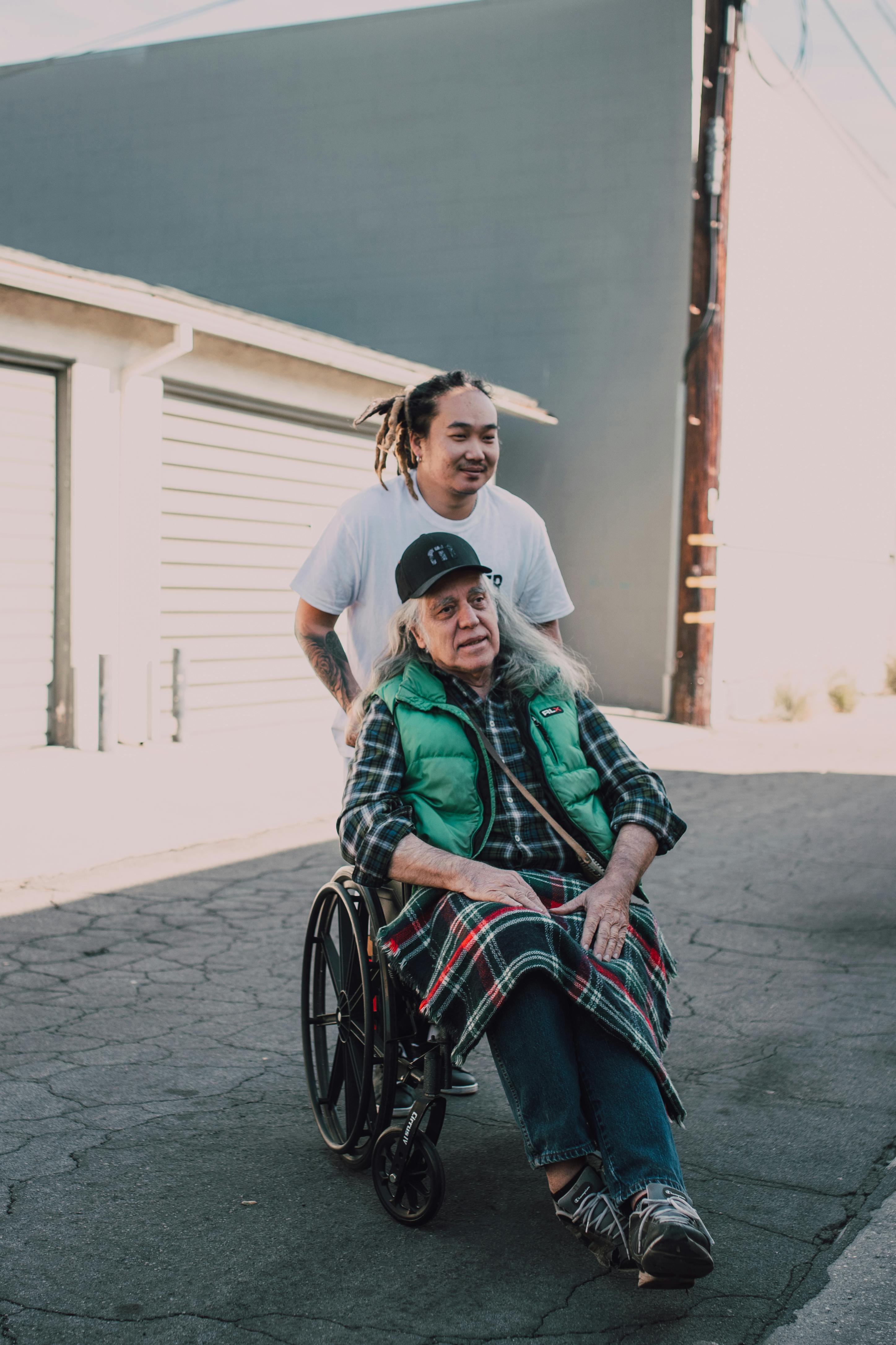Younger man with dreadlocks pushing an older man with long gray hair in a wheelchair along a sunlit street.