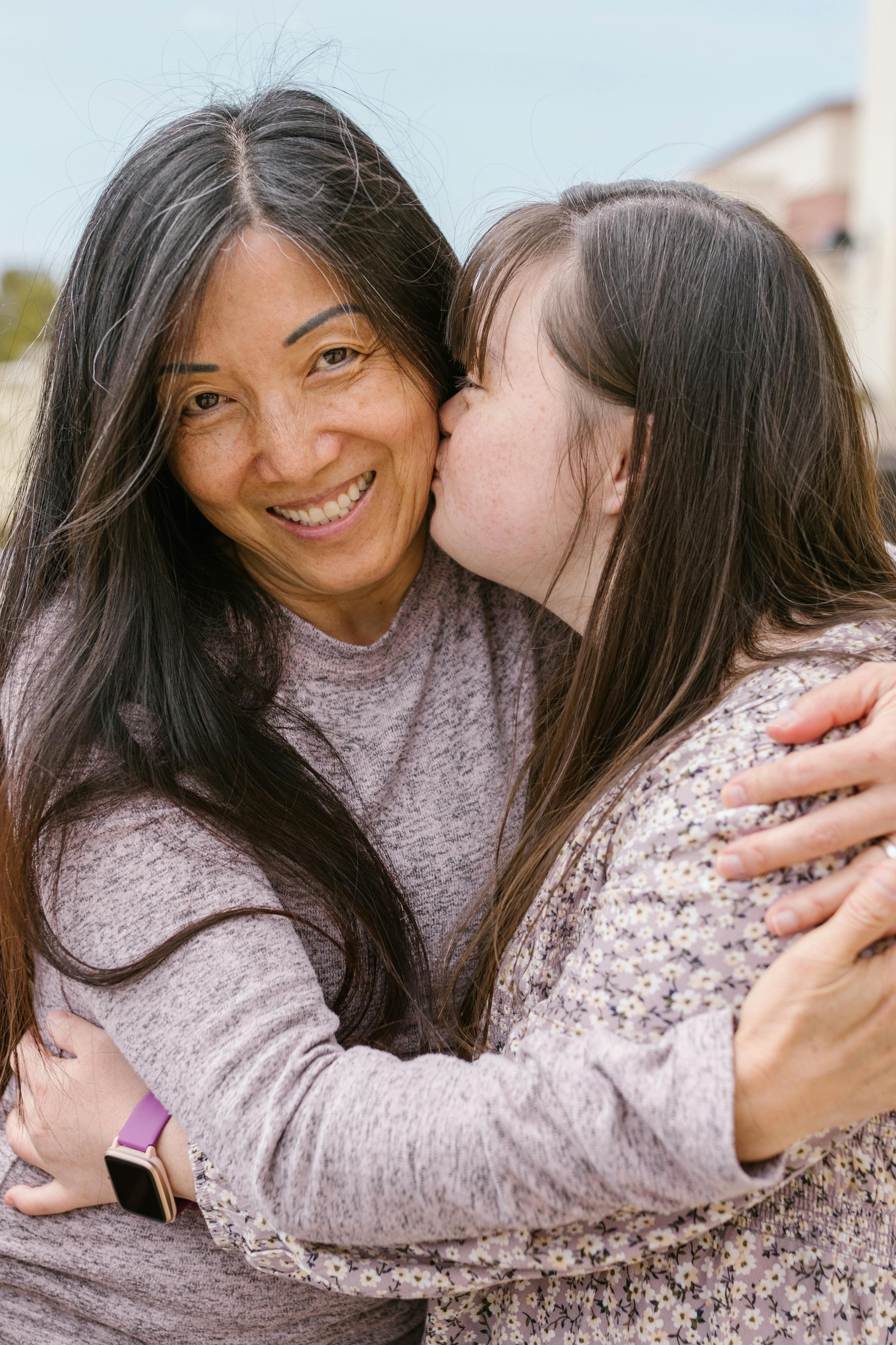 Young woman with Down syndrome kissing a smiling older woman on the cheek while hugging outdoors.