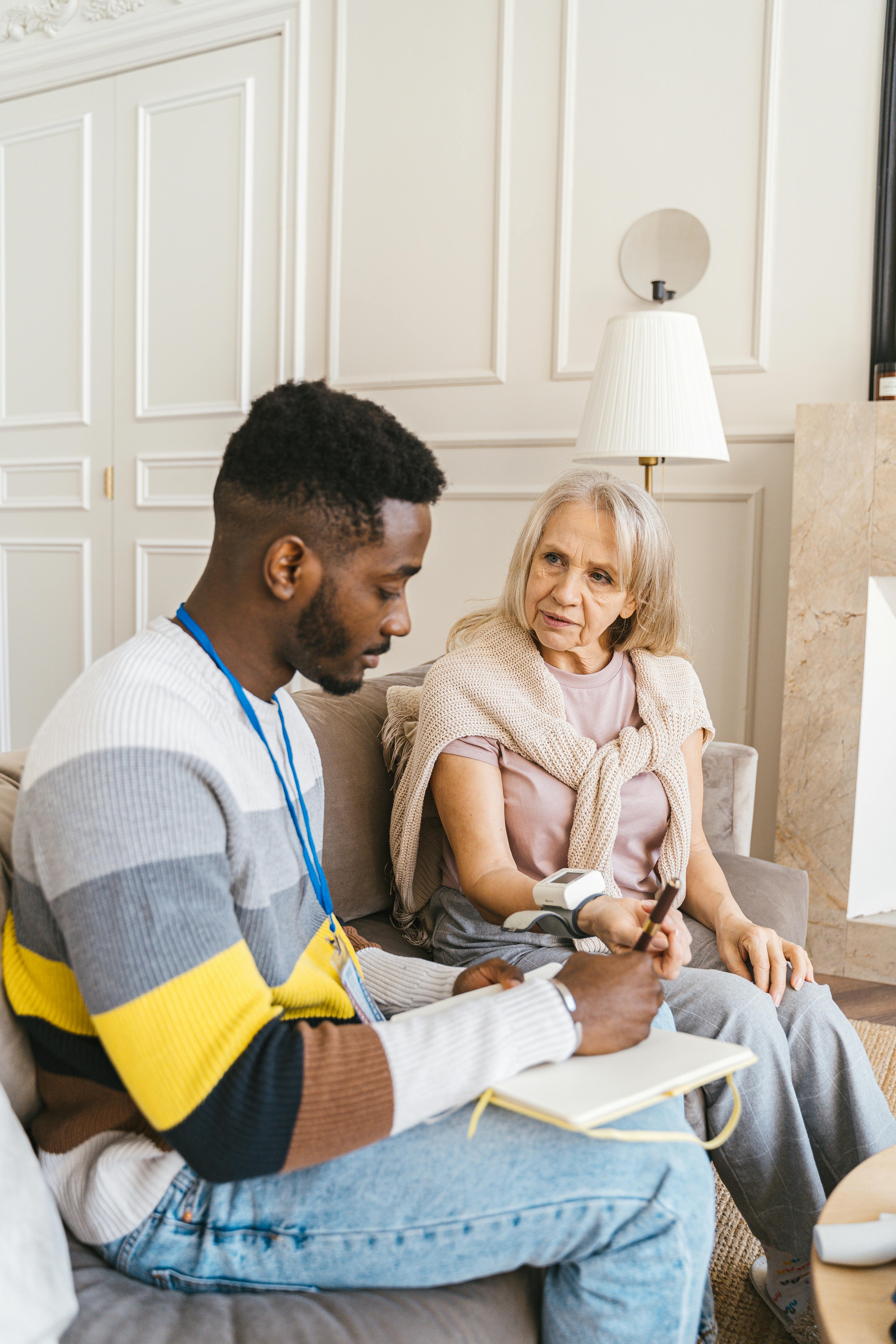 Young man in a striped sweater writing notes while checking blood pressure of an elderly woman seated on a couch in a living room.