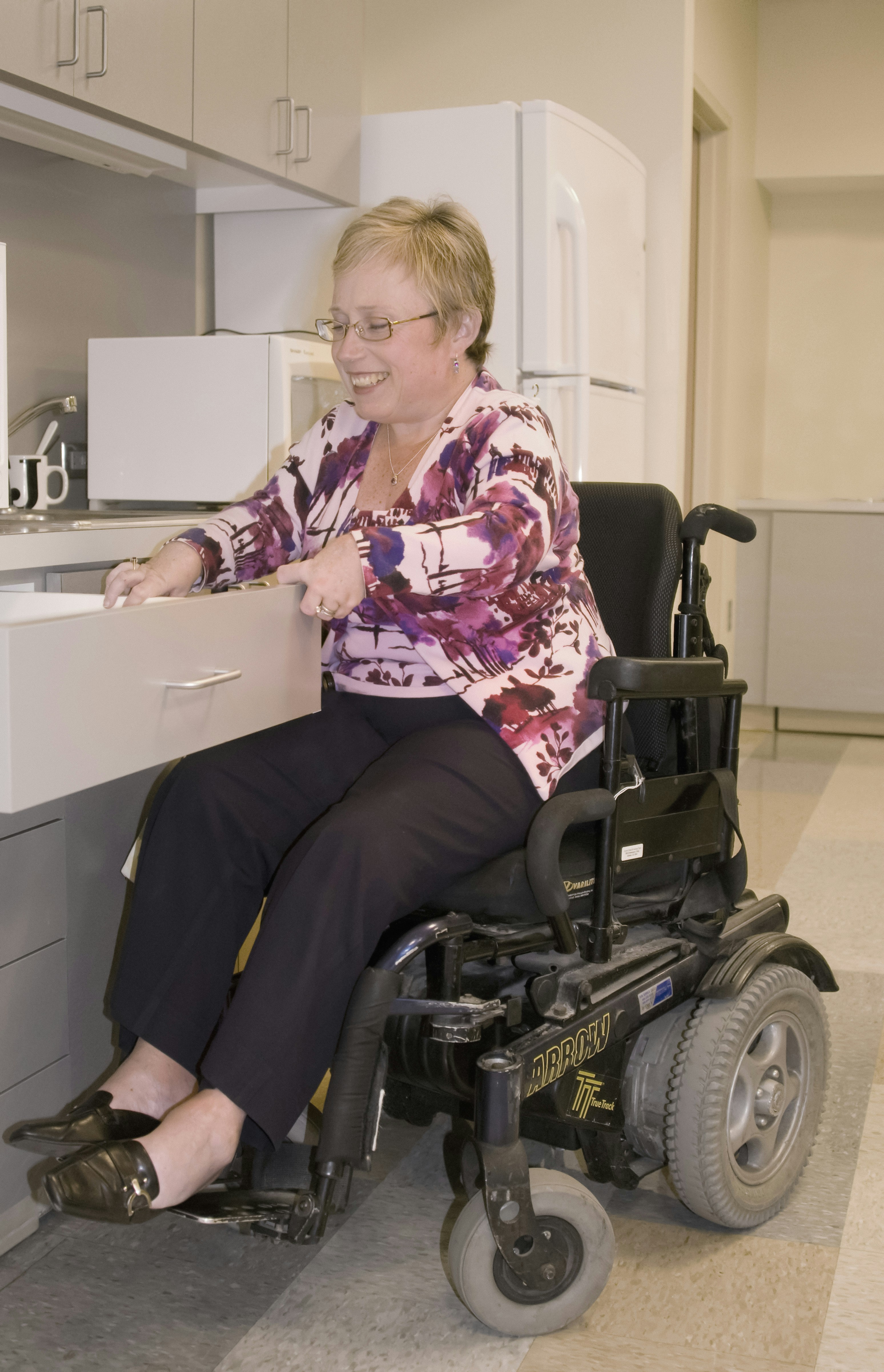 Smiling woman in a motorized wheelchair opening a kitchen drawer in a home setting.