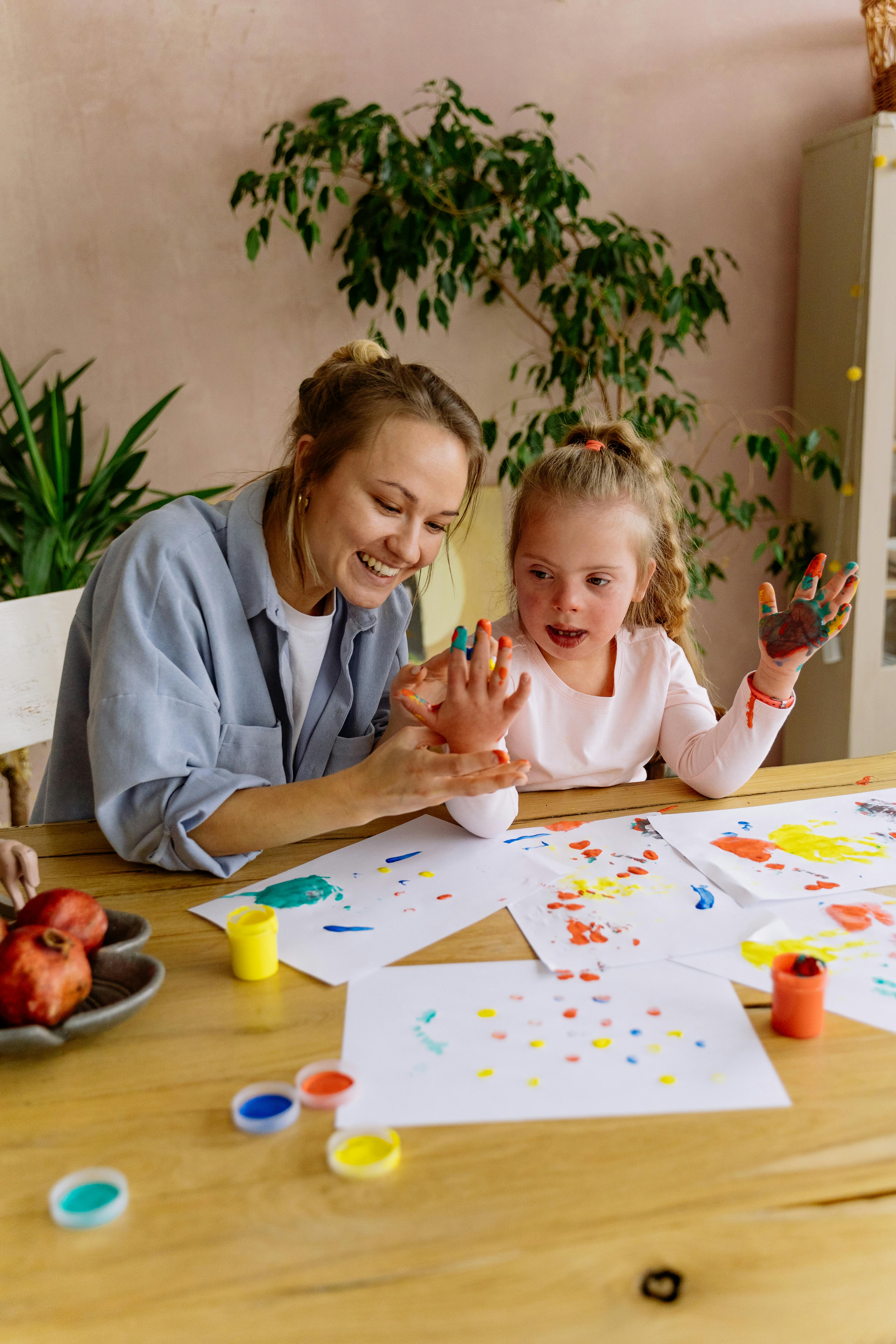 Smiling woman helps young girl with Down syndrome paint using colorful handprints at a wooden table.