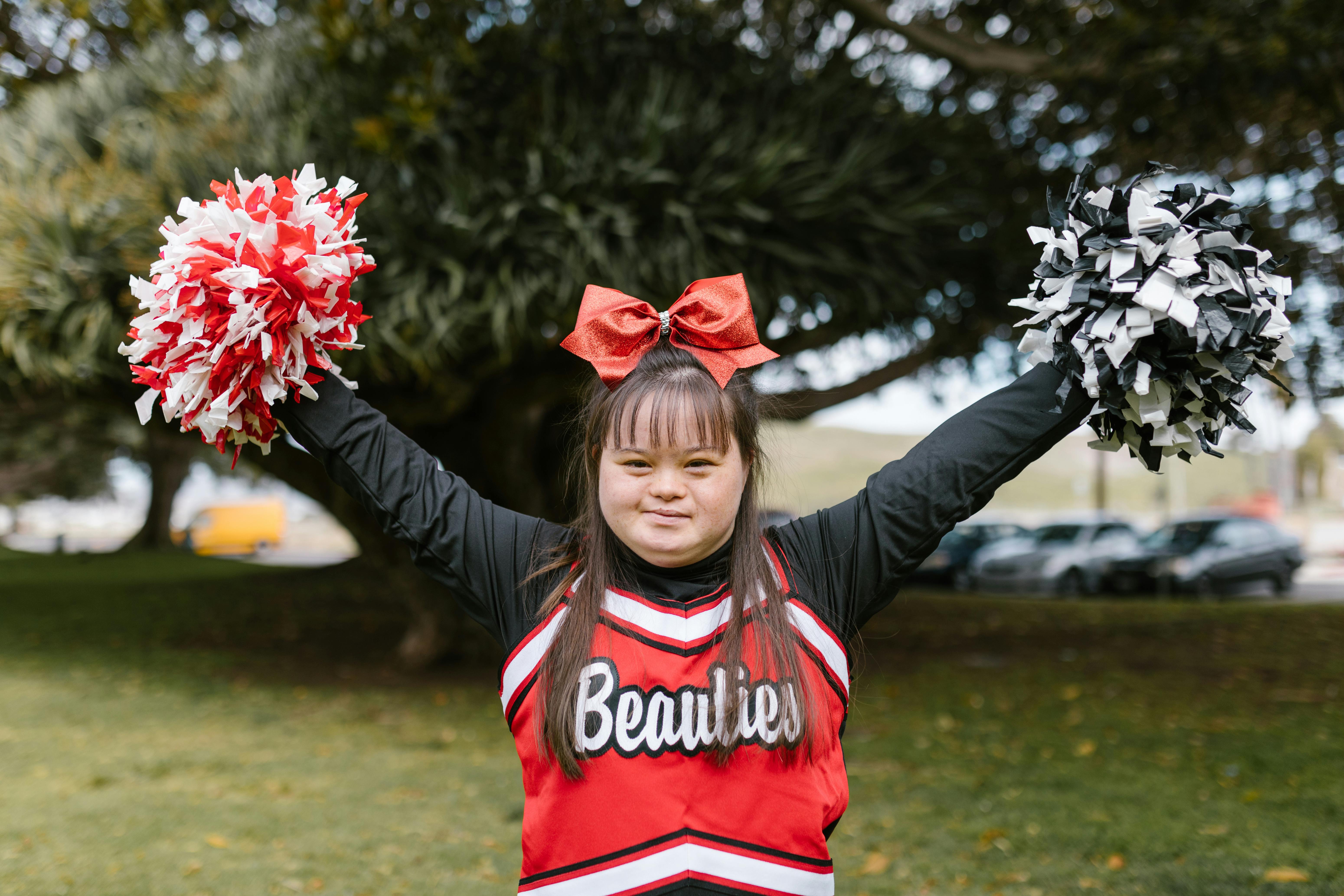 Cheerleader in red and black uniform with red bow and pom-poms raised outdoors.