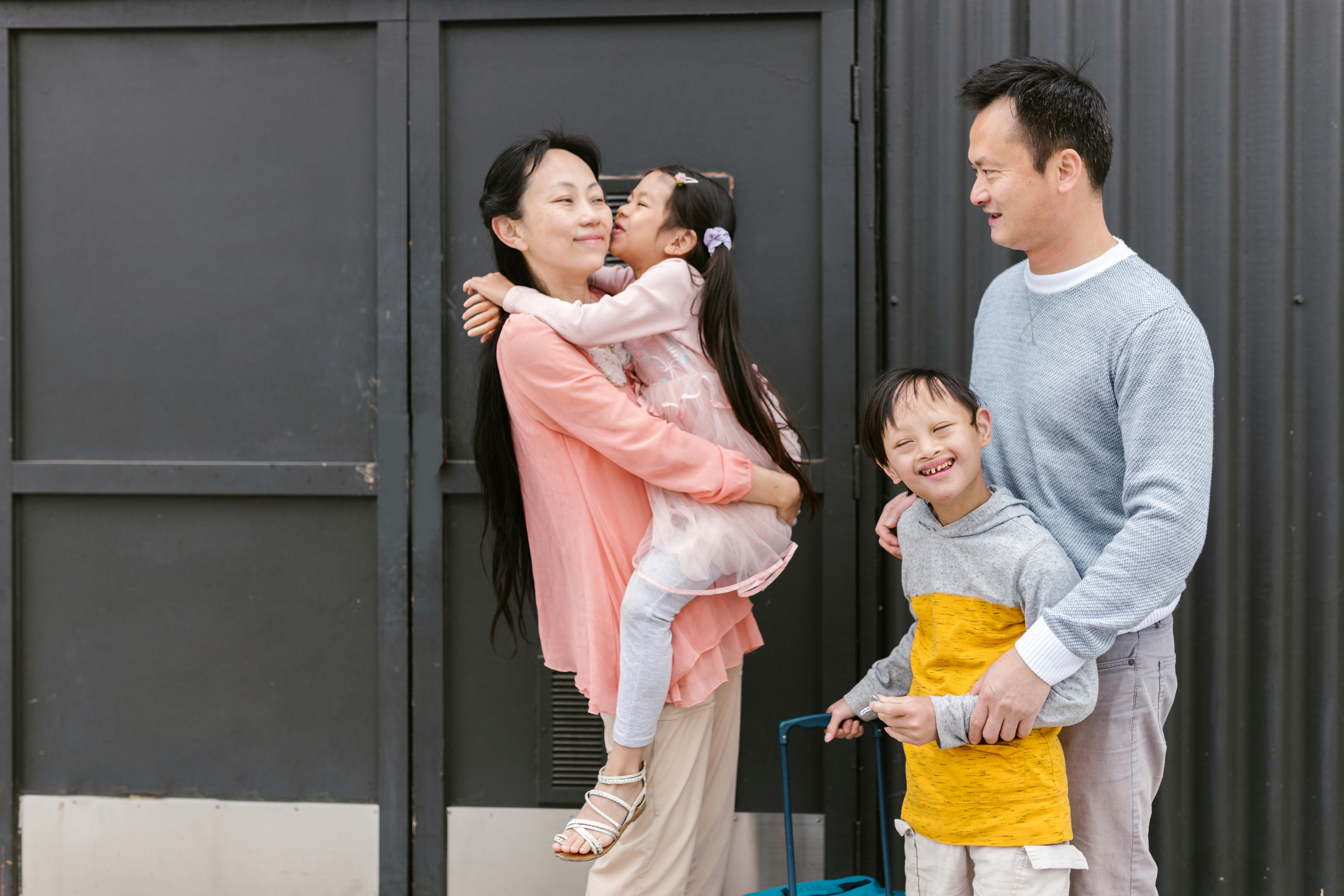 Asian family with mother holding daughter hugging her, father standing with son holding a suitcase.