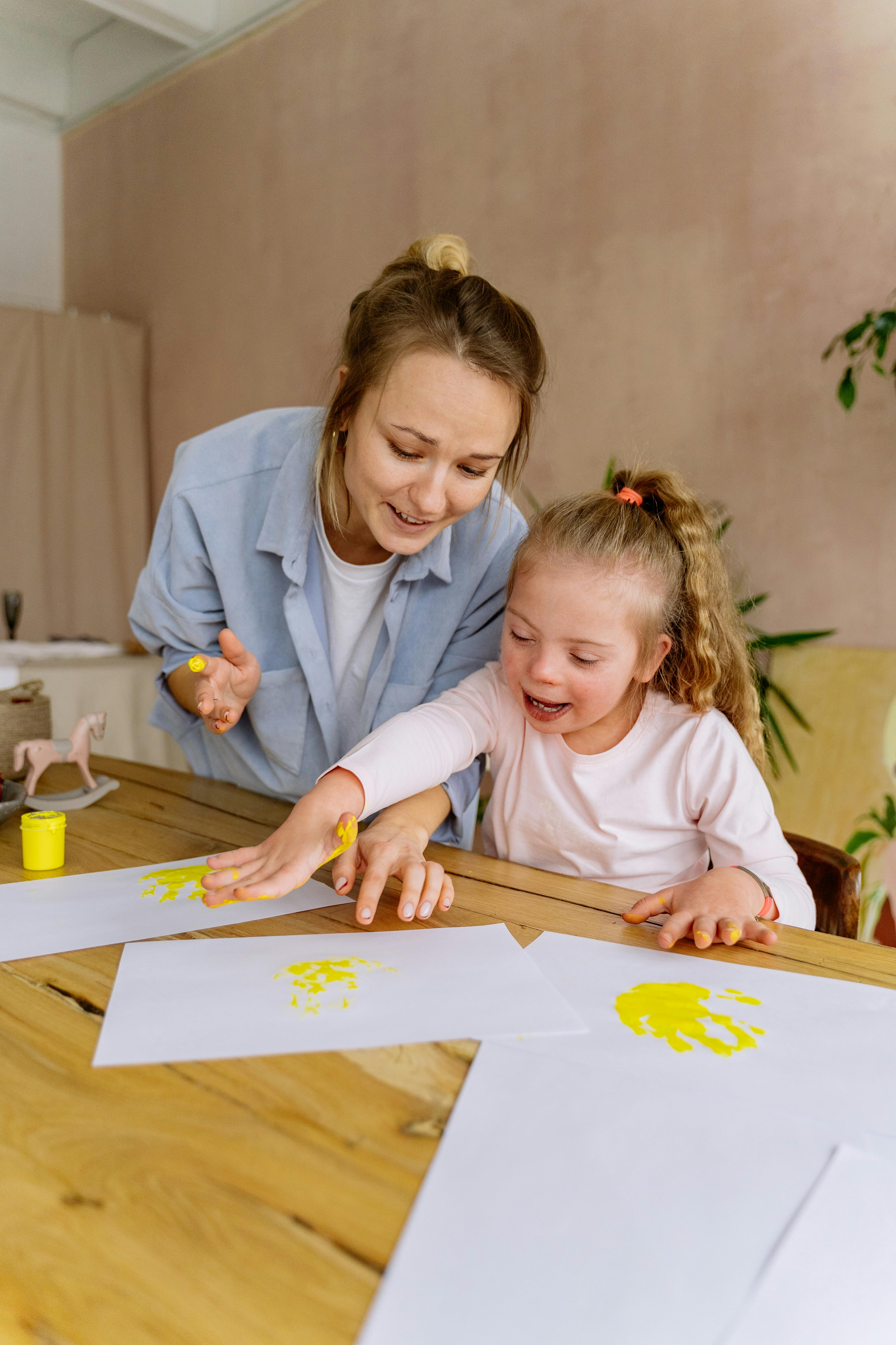 Woman and young girl happily making yellow handprints on paper together at a wooden table.