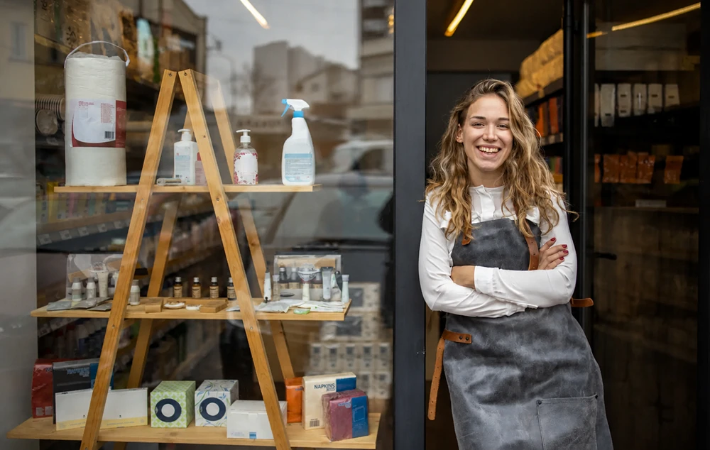 Jeune femme souriante portant un tablier debout à la porte d'une boutique avec un panneau ouvert.