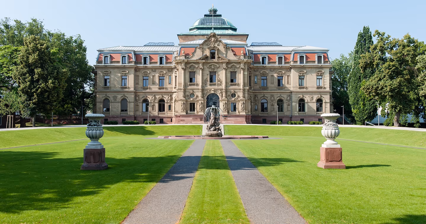 Repräsentatives historisches Gerichtsgebäude (Bundesgerichtshof) mit symmetrischer Fassade und Kuppeldach; davor ein gepflegter Rasen mit geradem Weg und einem Brunnen/Skulptur in der Mitte – Symbolbild für Justiz und Rechtsprechung.