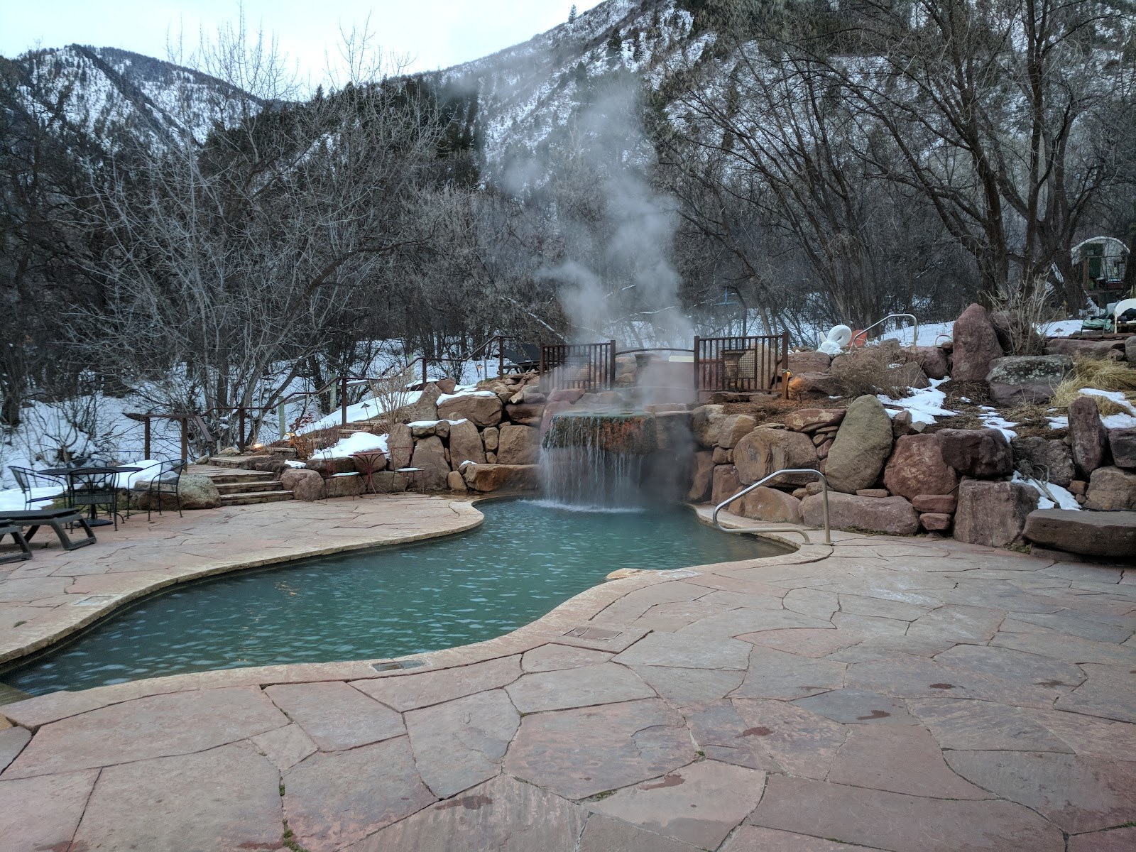 Steaming outdoor hot spring pool with a waterfall and snowy mountain backdrop at Avalanche Ranch Cabins & Hot Springs in Colorado.