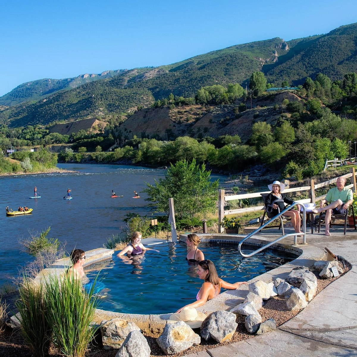 People relaxing in an outdoor hot spring pool by a river at Iron Mountain Hot Springs near Glenwood Springs, Colorado on a sunny day