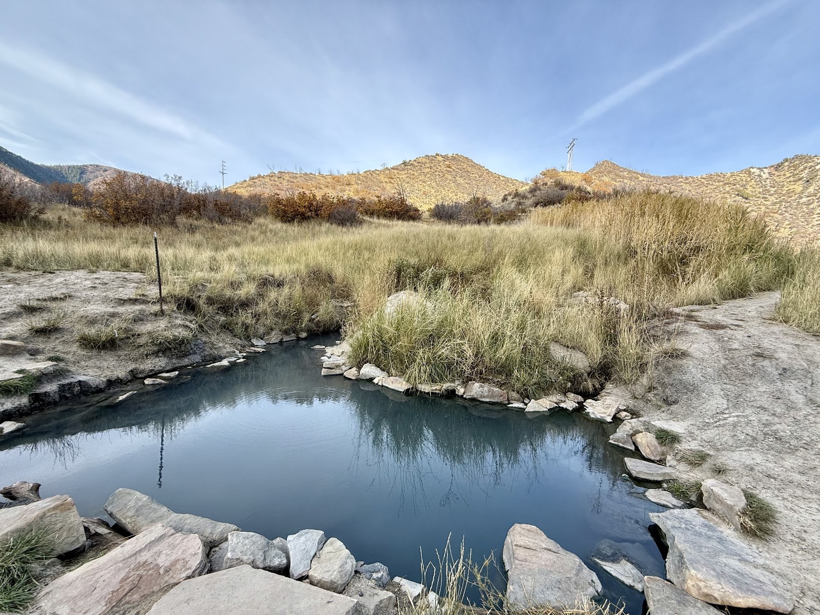 Calm pool of South Canyon Hot Springs bordered by stones and dry grass with hills in the background near Glenwood Springs, Colorado.