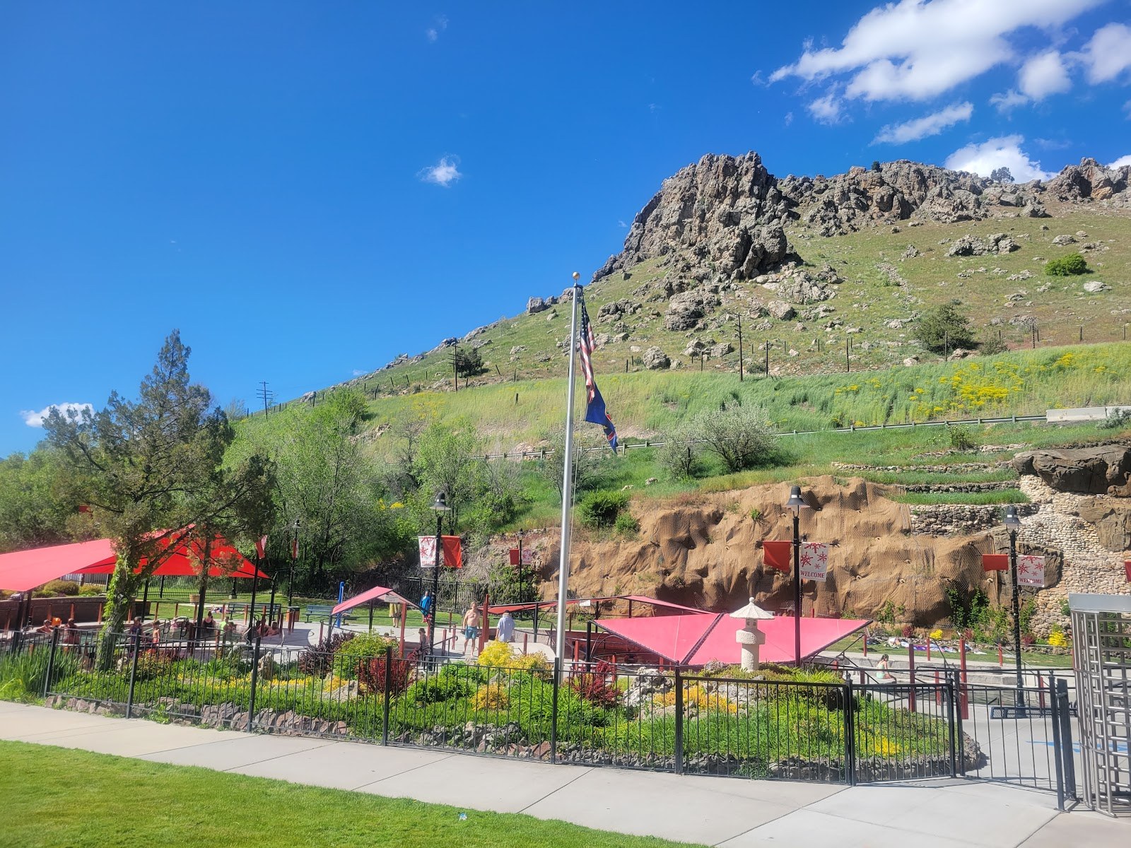 Daytime wide-angle view of Aura Soma Lava Hot Springs in Lava Hot Springs, Idaho, with vibrant red canopies, green landscape, and rocky mountain backdrop.