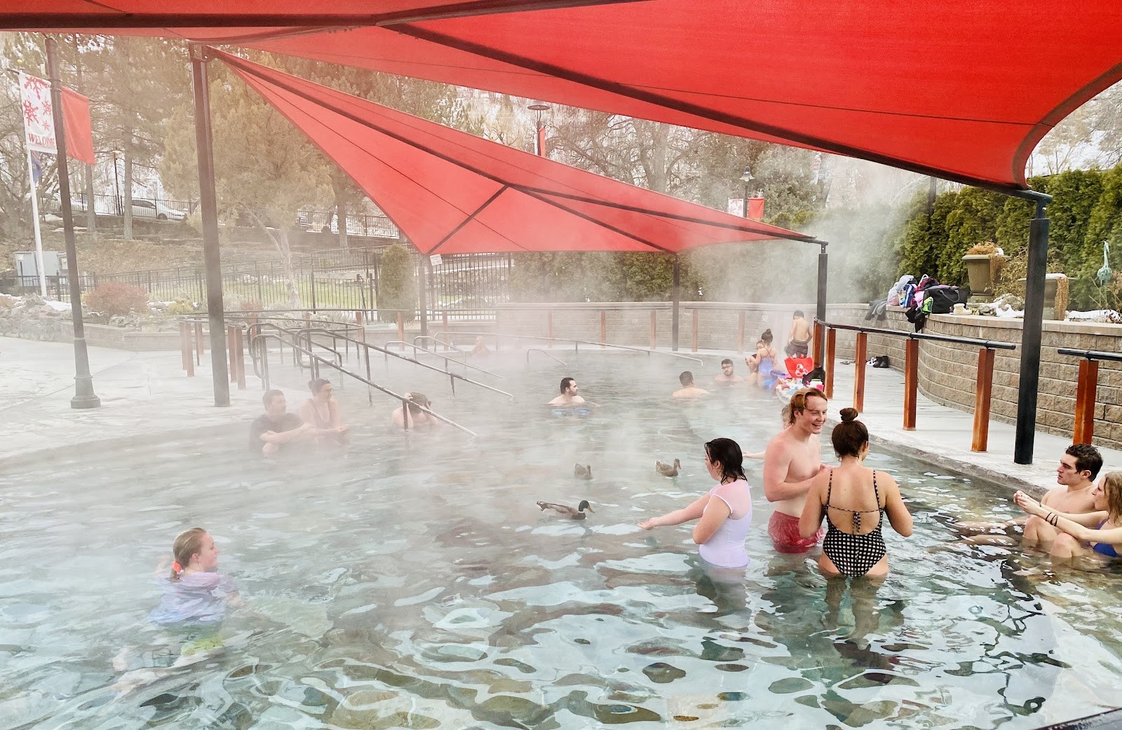 Outdoor hot spring pool at Aura Soma Lava Hot Springs in Lava Hot Springs, Idaho, with people relaxing under red canopies and visible steam.