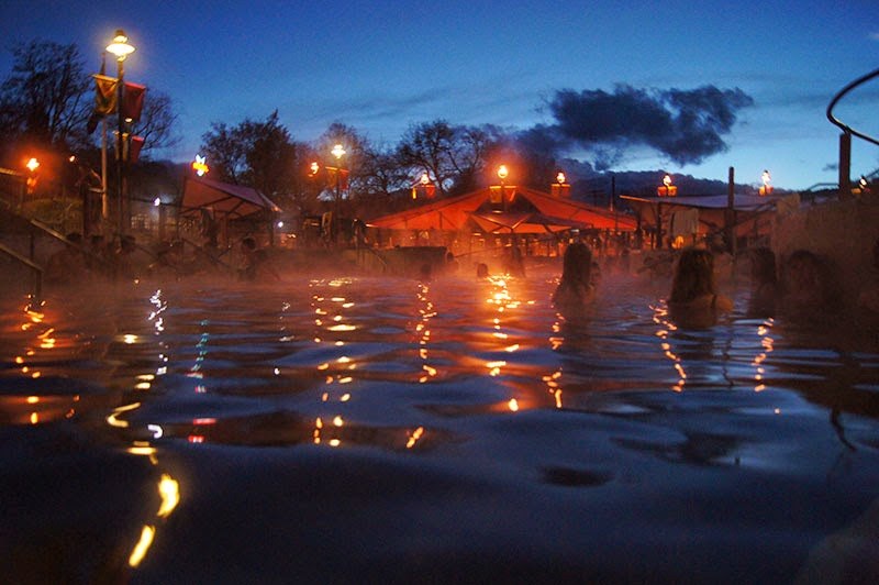 Evening view of Aura Soma Lava Hot Springs in Lava Hot Springs, Idaho, showing people enjoying the steamy hot spring pool with warm lights reflecting on water.