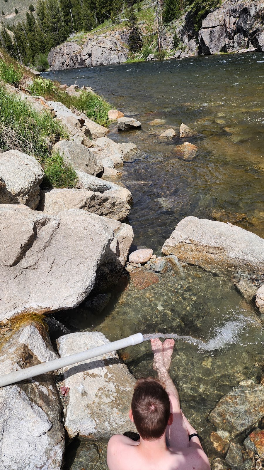 Clear water of Boat Box Hot Spring near a rocky riverbank in Stanley, Idaho, with green vegetation and forested hills