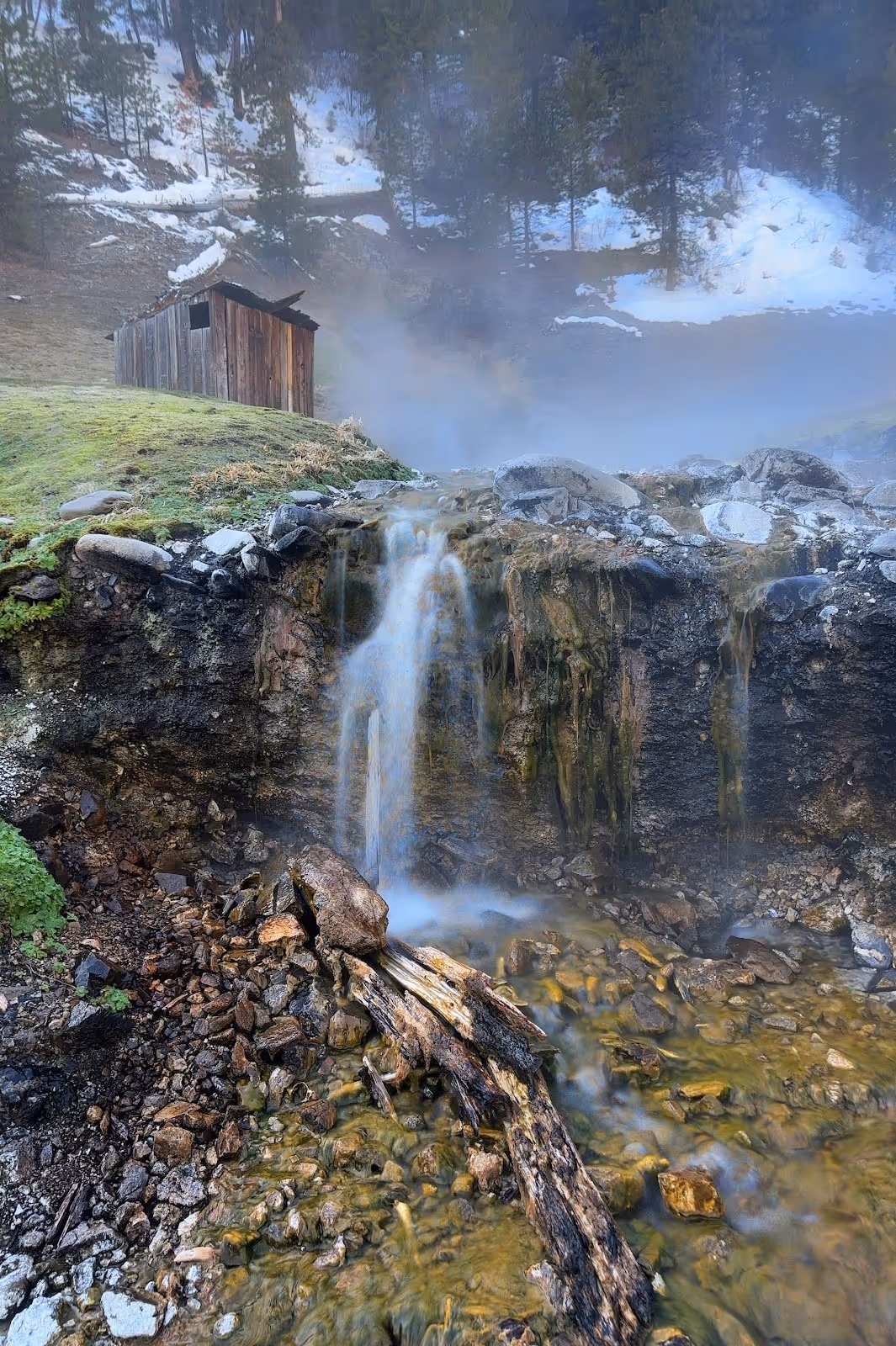 Waterfall flowing from rocky terrain near an old wooden cabin with steam rising at Bonneville Hot Springs in Lowman, Idaho, United States