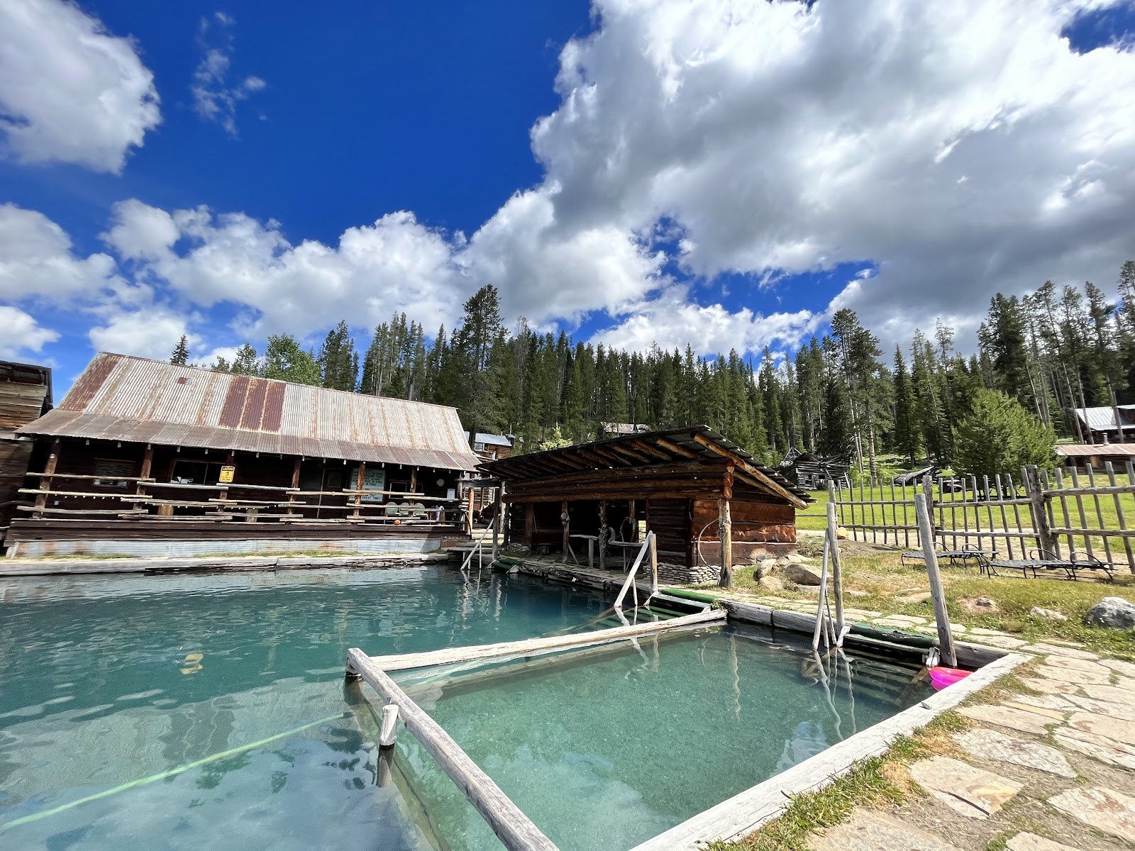 Clear outdoor hot spring pool with rustic wooden structures under a blue sky at Burgdorf Hot Springs near McCall, Idaho, United States.