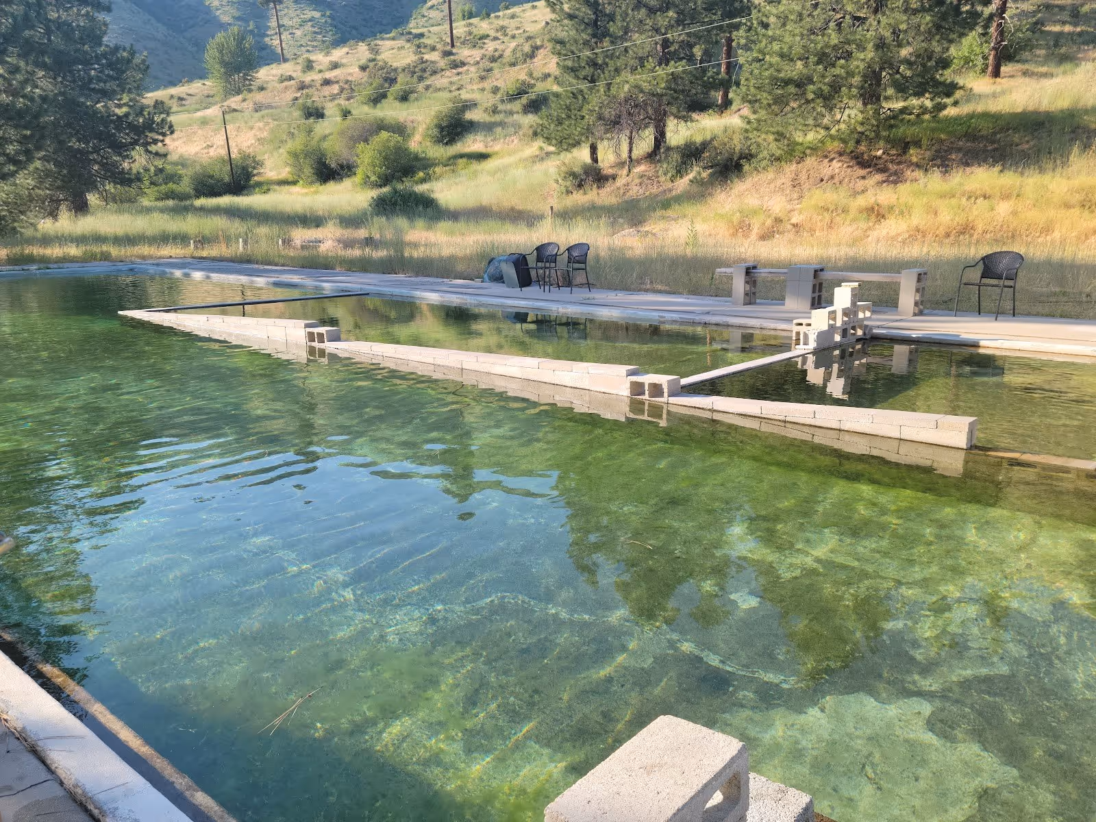 Clear water of Haven Hot Springs outdoor pool in Lowman, Idaho, surrounded by green hills and a few chairs along the concrete edge.