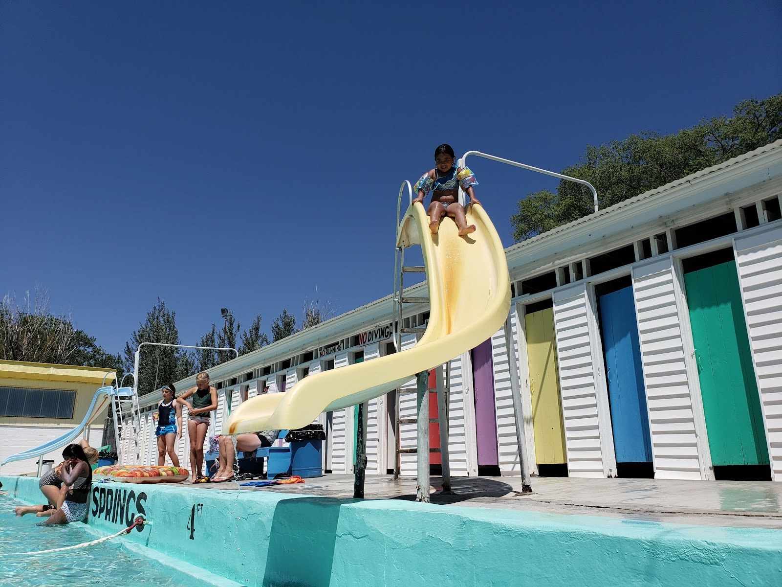 A child sliding down a yellow slide into the outdoor hot spring pool at Indian Springs Resort with colorful changing cabins in American Falls, Idaho.