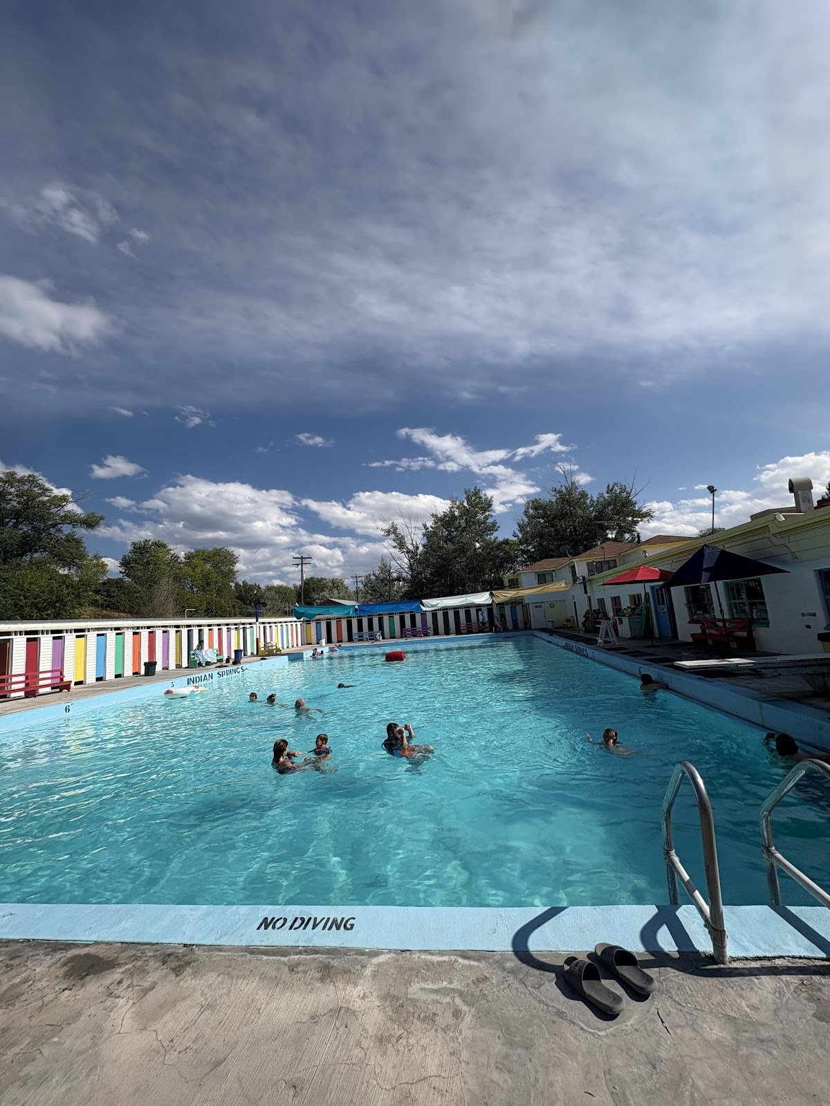 People enjoying swimming in the outdoor hot spring pool of Indian Springs Resort near American Falls, Idaho, under a partly cloudy sky.